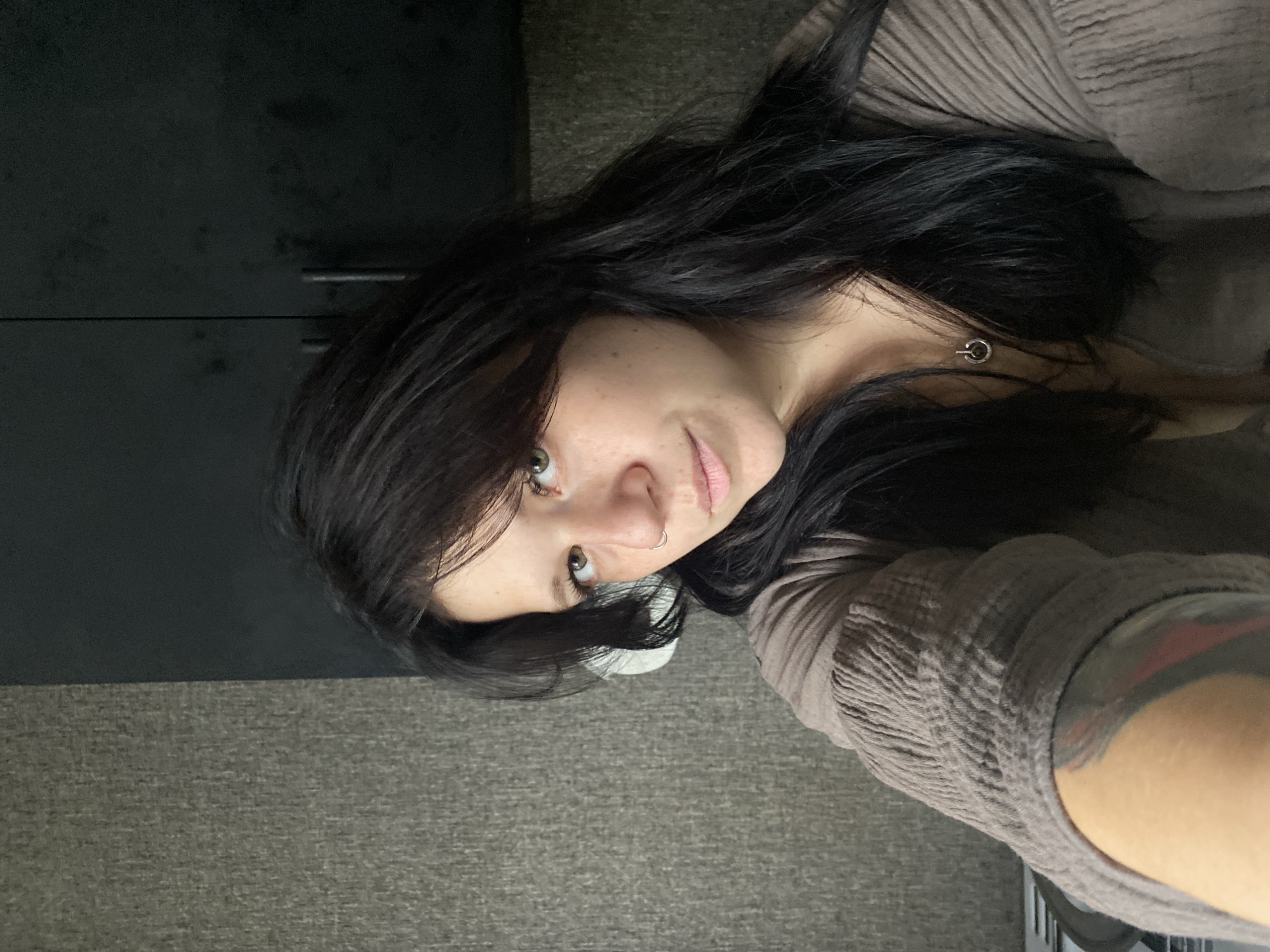 A medium shot shows a woman with dark hair and light skin, looking directly at the camera. She is wearing a brown, textured top and a silver necklace. Her left arm is visible, displaying a tattoo. The background is divided into two sections: a textured gray wall on the left and a dark, flat surface on the right, possibly cabinetry. The lighting is soft, casting gentle shadows. The scene appears to be indoors, in a residential setting. No specific location in Chaikovsky, Russia is identifiable.