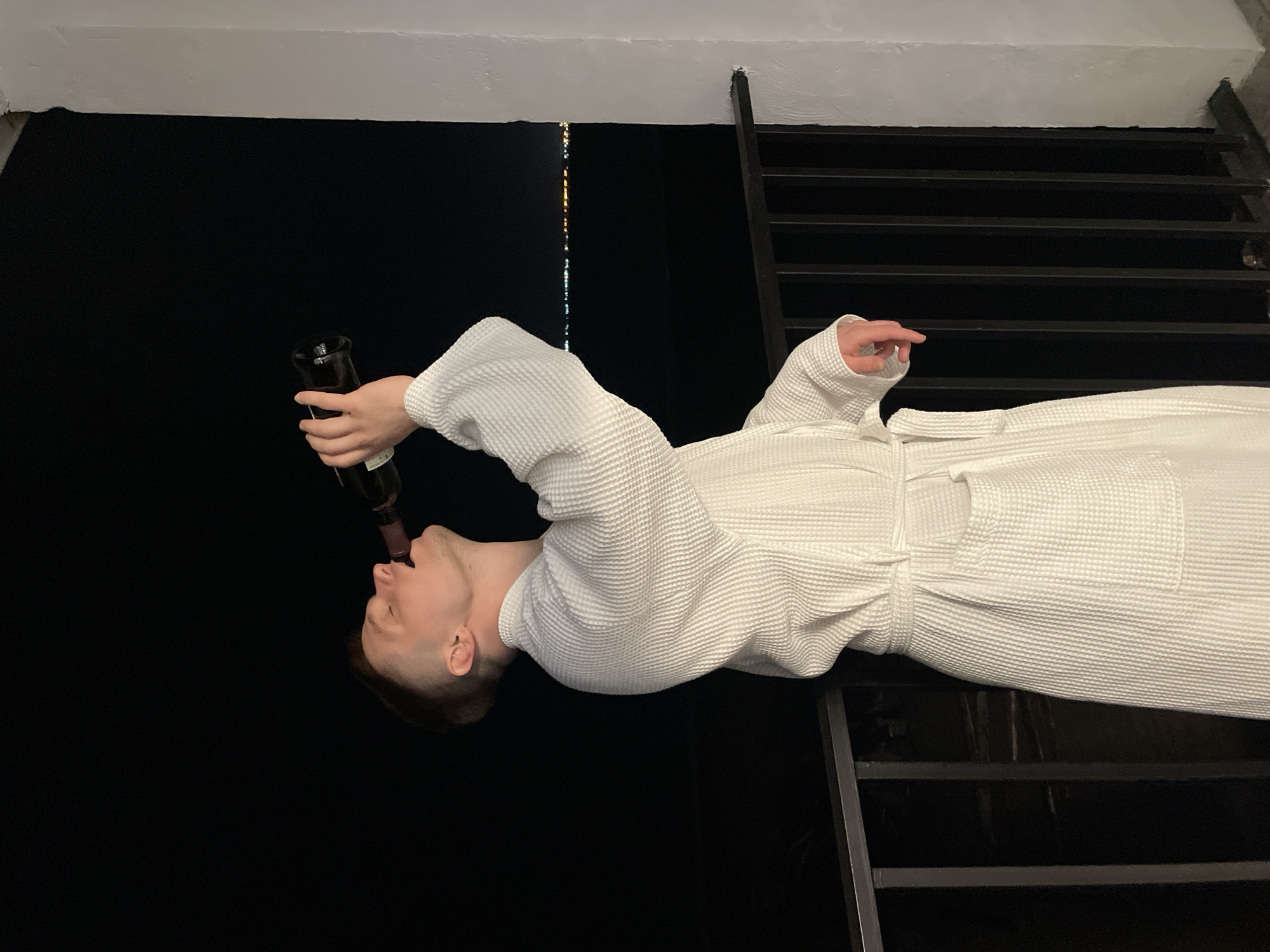 A person wearing a white waffle-weave bathrobe is drinking directly from a dark-colored bottle. The background appears to be a night scene, possibly outdoors, with dark or blacked-out areas. A black metal ladder is visible on the right side of the frame. The location is Unknown, Unknown.