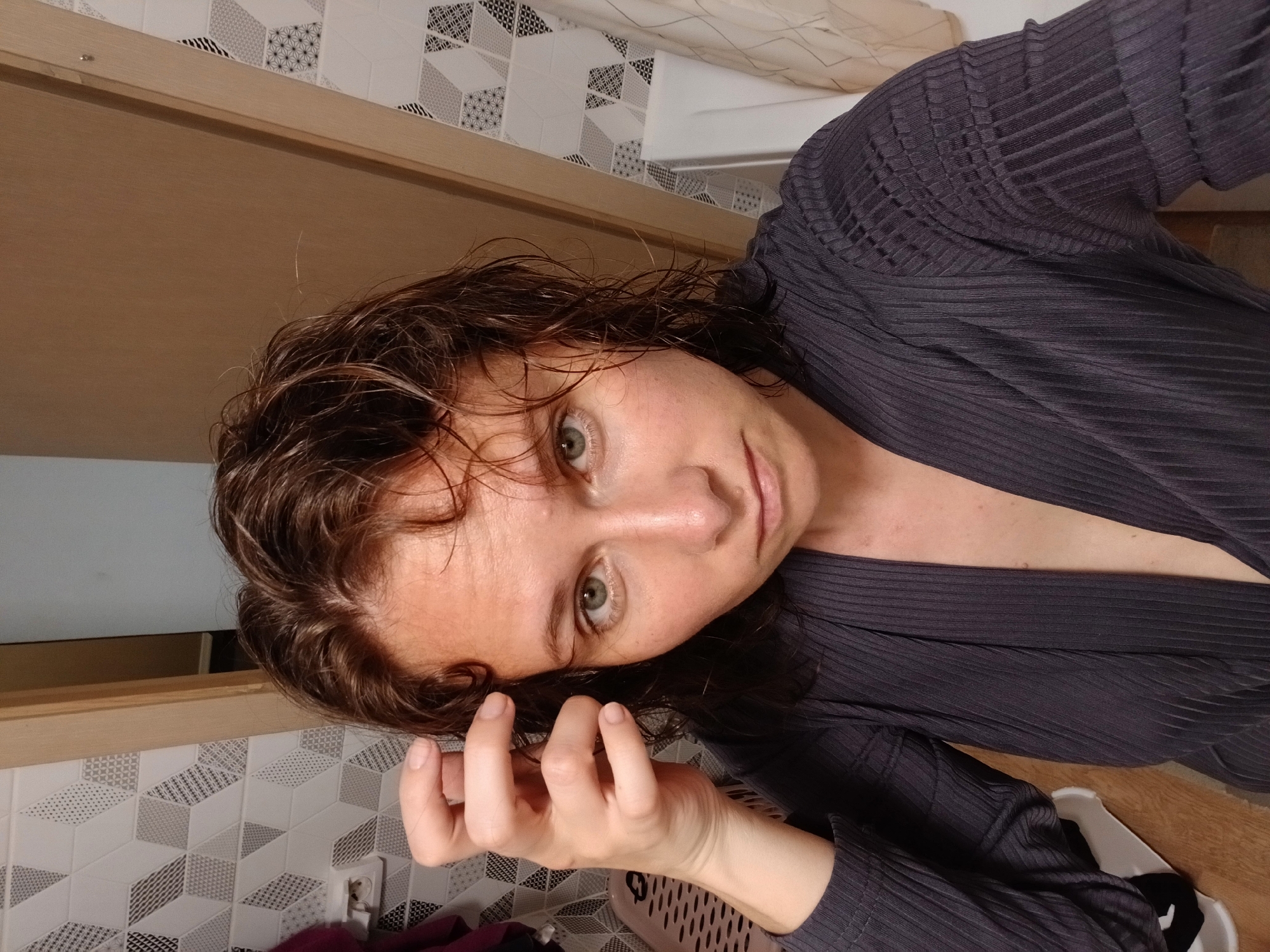 The media file is a selfie taken by a young woman indoors. She has dark, wavy hair that appears wet, and light green eyes. She is wearing a dark grey, ribbed robe or cardigan. Her left hand is resting near her face, touching her hair. The background shows a wall with patterned tiles, a wooden door frame, and a glimpse of a laundry basket. The lighting suggests it could be daytime. The overall mood is casual and relaxed. There is a visible electrical outlet on the tiled wall. The location context is Yekaterinburg, Russia.