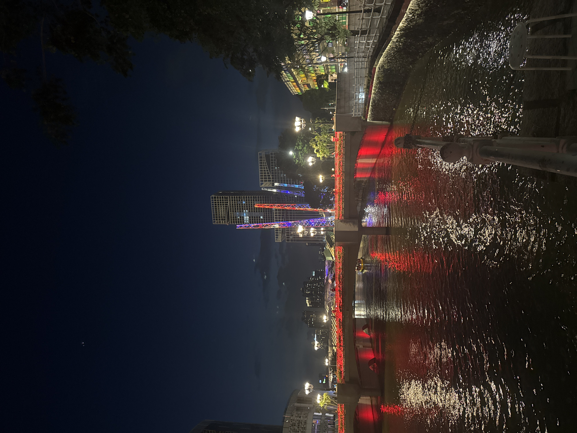 The image depicts a night scene along the Singapore River in Singapore. A bridge illuminated with red lights spans the river, reflecting brightly on the water's surface. On the bridge, figures can be seen walking and enjoying the view, with additional red lights lining the sides.
In the background, modern skyscrapers dominate the skyline, some adorned with colorful lights, creating a vibrant urban panorama. The riverbanks are lined with buildings and trees, further enhancing the cityscape. The water itself is a dark, reflective surface, mirroring the lights from the bridge and surrounding structures.
The sky above is a deep, dark blue, with a few faint clouds visible. Two small, bright points of light, possibly stars or planets, are discernible in the upper left portion of the sky. The overall atmosphere is one of a lively urban night, characterized by illuminated architecture and the tranquil flow of the river.