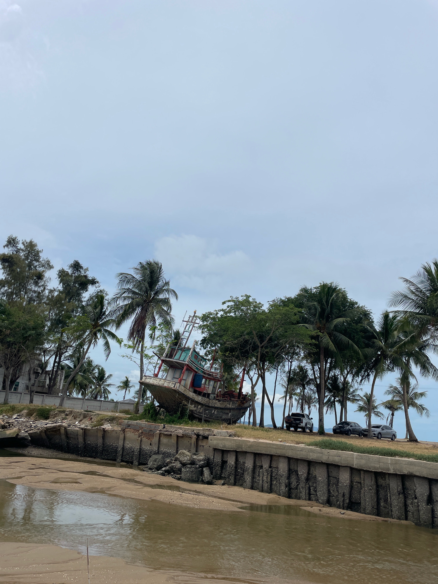 The image depicts a weathered, beached fishing boat on the shore of a body of water, likely a canal or inlet, in Ban Na Chom Thian, Thailand. The boat is tilted on its side, revealing its hull and deck, with remnants of its structure and some debris visible. Coconut palm trees and other lush green vegetation line the shore behind the boat, with a concrete retaining wall separating the vegetation from the water. Several vehicles, including a pickup truck and two sedans, are parked on the grassy area above the retaining wall, near the trees. The sky is overcast, suggesting a cloudy day. The water in the foreground is calm and appears murky. The overall scene conveys a sense of decay and abandonment, juxtaposed with the tropical environment.