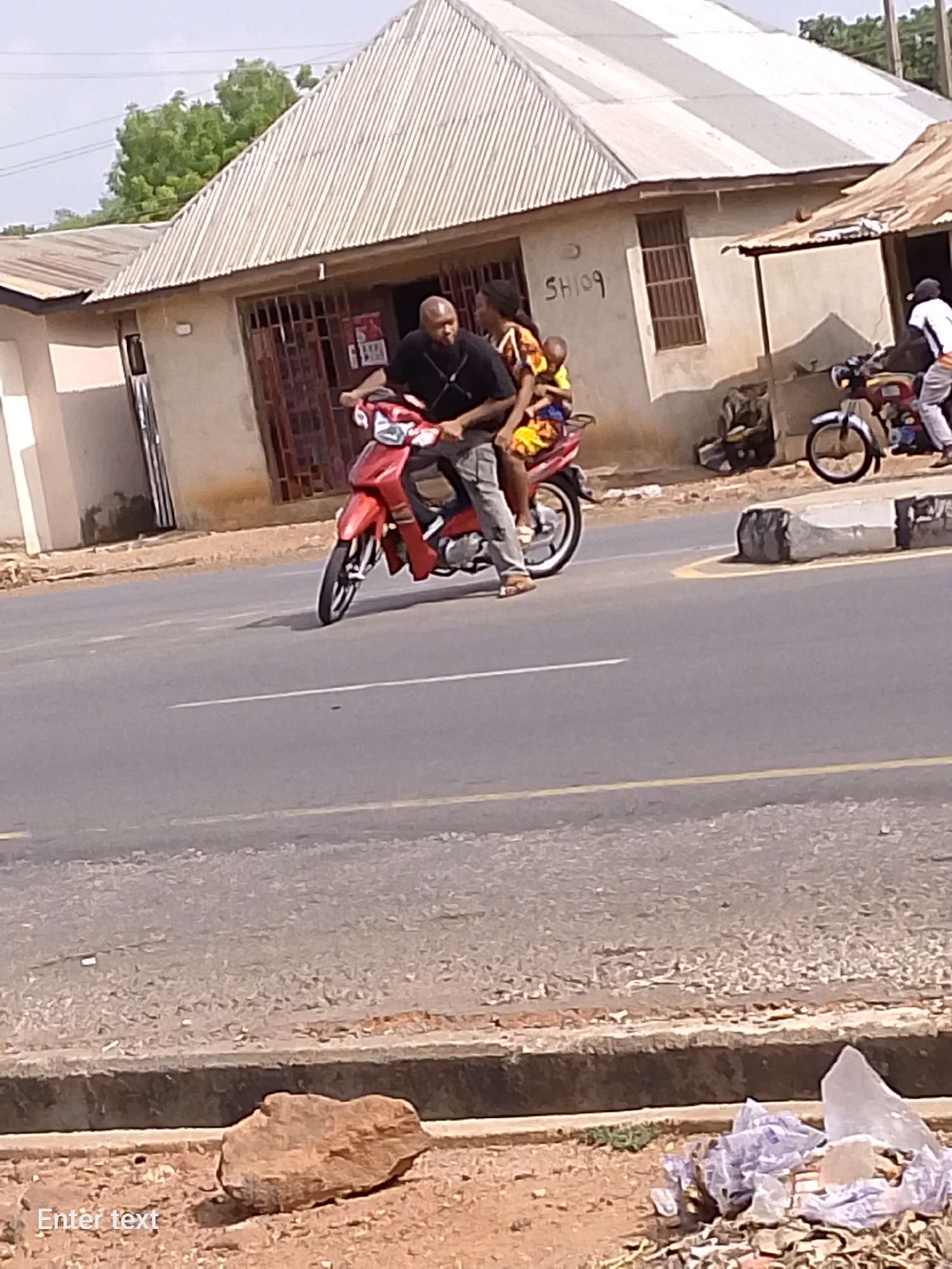 A man is riding a red motorcycle with a woman and child as passengers. The location is Adankolo, Nigeria. The riders are on a paved road with buildings on either side. Other motorcycles and people are visible in the background. There is trash along the curb.