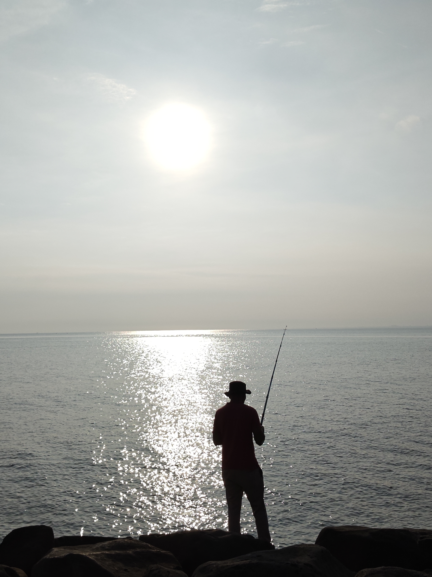 An outdoor vertical shot captures a lone individual fishing at the coast in Lhokseumawe, Indonesia. The person, viewed from behind, is rendered in dark silhouette against the bright backdrop of the sky and sea. They stand on large, dark rocks at the water's edge, wearing a hat and holding a fishing rod extended towards the calm ocean.
The sun is a brilliant, almost perfectly round light source, positioned in the upper left of the frame. Its intense light creates a dazzling, sparkling path across the water's surface, stretching from the horizon towards the foreground. The sky is a hazy, light blue-grey with minimal, indistinct clouds. The strong backlighting makes the distant horizon appear blended with the sky, adding to the serene atmosphere.
The scene conveys a sense of peaceful solitude, with the person engaged in the quiet activity of fishing. The sun's low position and strong reflection suggest it is either early morning or late afternoon. No visible text is present in the image.