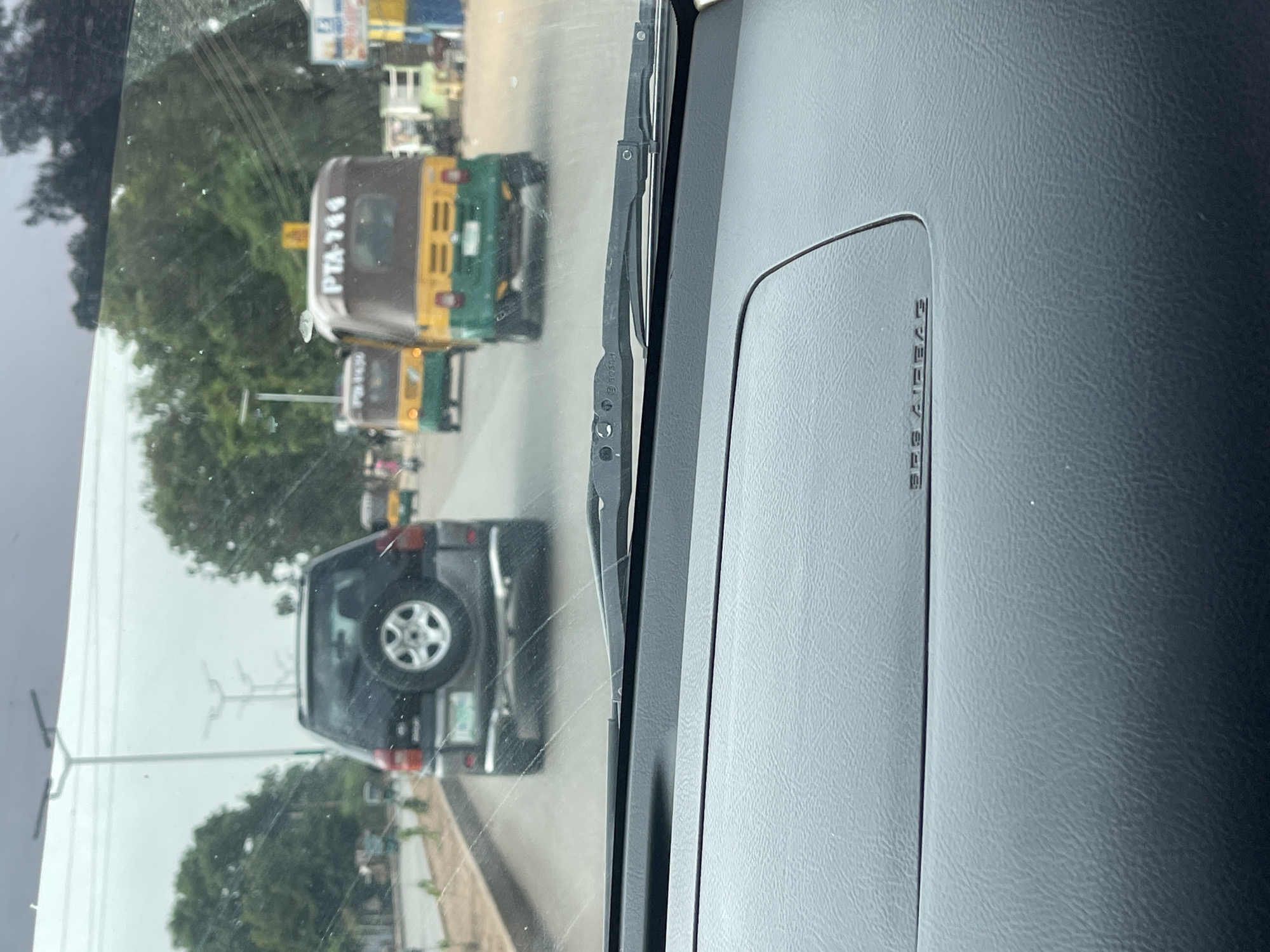 The image is taken from inside a vehicle, looking out through the windshield at a street scene. In front of the vehicle is a dark-colored SUV with a spare tire mounted on its rear. Further ahead are two yellow and green "Keke Napep" tricycles, one with the registration "PTA-744." The setting appears to be urban, with trees and buildings visible along the roadside. Given the vehicle is in Jos, Nigeria and the mention of "Keke Napep," it is highly likely the location is in Jos, Nigeria. The sky is overcast, suggesting a cloudy day. The interior of the vehicle shows a dashboard with the inscription "SRS AIRBAG." A windshield wiper is partially visible in the center of the frame.