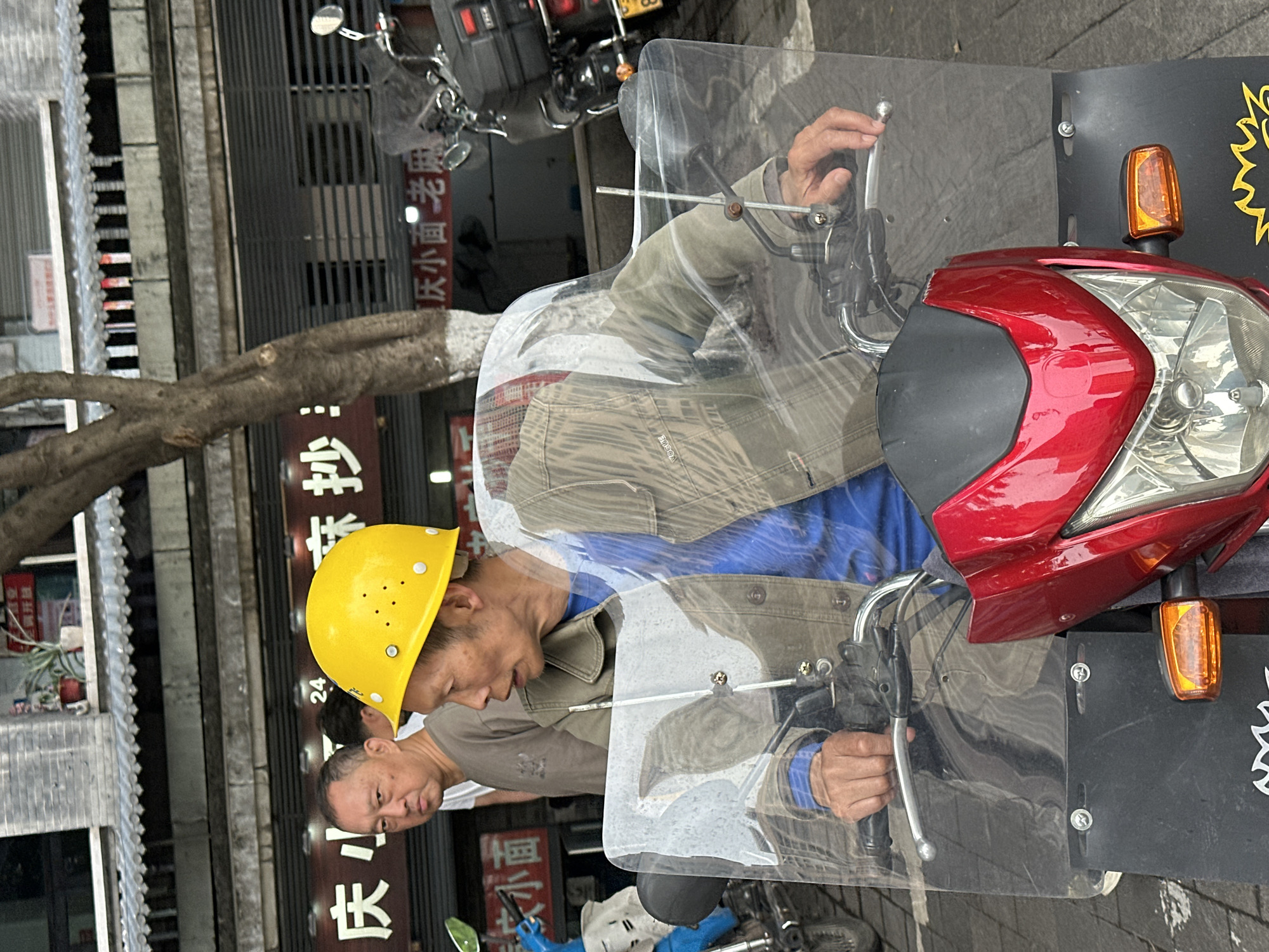 The image shows a man wearing a yellow construction helmet riding a red motorcycle, possibly in Shapingba District, China. He is wearing a blue shirt and a khaki jacket. The motorcycle has a clear windshield and a black back. The background shows buildings with Chinese characters. There is a tree to the left of the buildings. A second man, possibly the rider's companion, is visible.
