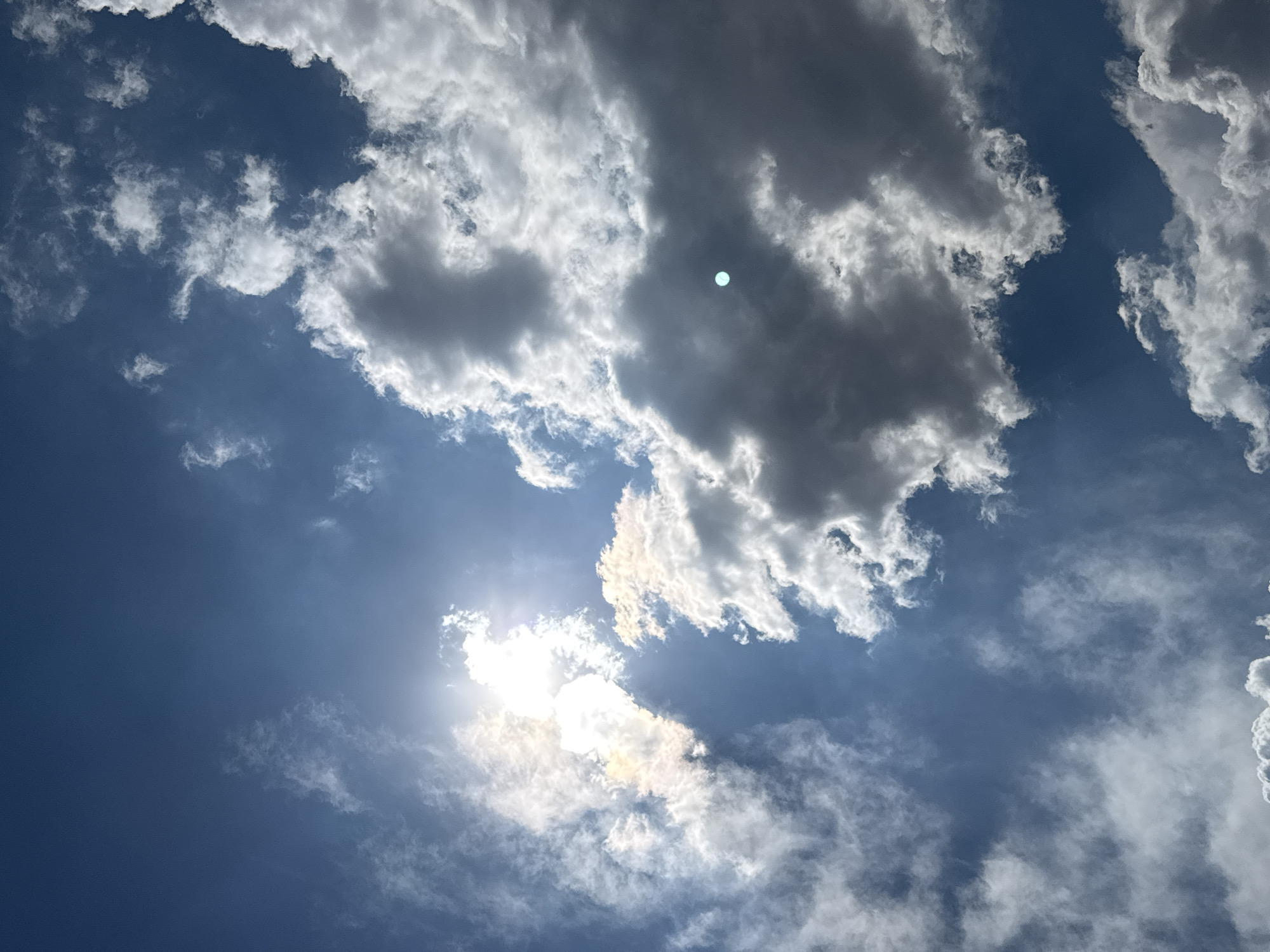 The image shows a view of the sky in San Francisco Ocotlán (Ocotlán), Mexico. The sky is predominantly blue with varying cloud formations. Some clouds are bright white, while others are darker gray. The sun is visible, partially obscured by clouds.