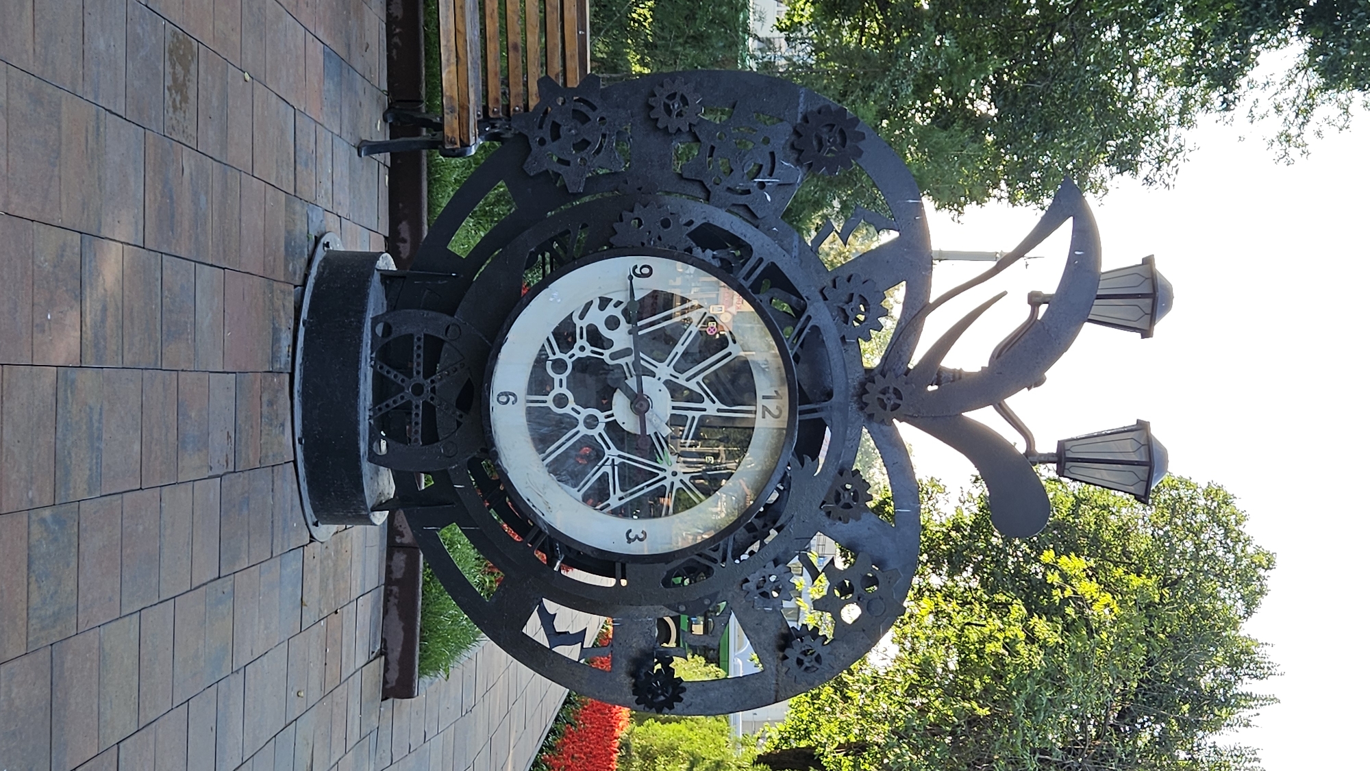 A large, ornate, black metal clock, designed to resemble an apple with gears, stands outdoors on a paved walkway. The clock face, with white Roman numerals, is visible through a glass cover. The clock is adorned with numerous metal gears of various sizes. Two decorative street lamps are mounted on top of the clock's structure. To the left of the clock is a wooden bench. The background features lush green trees and a vibrant flower bed. The scene appears to be in a park or public square.