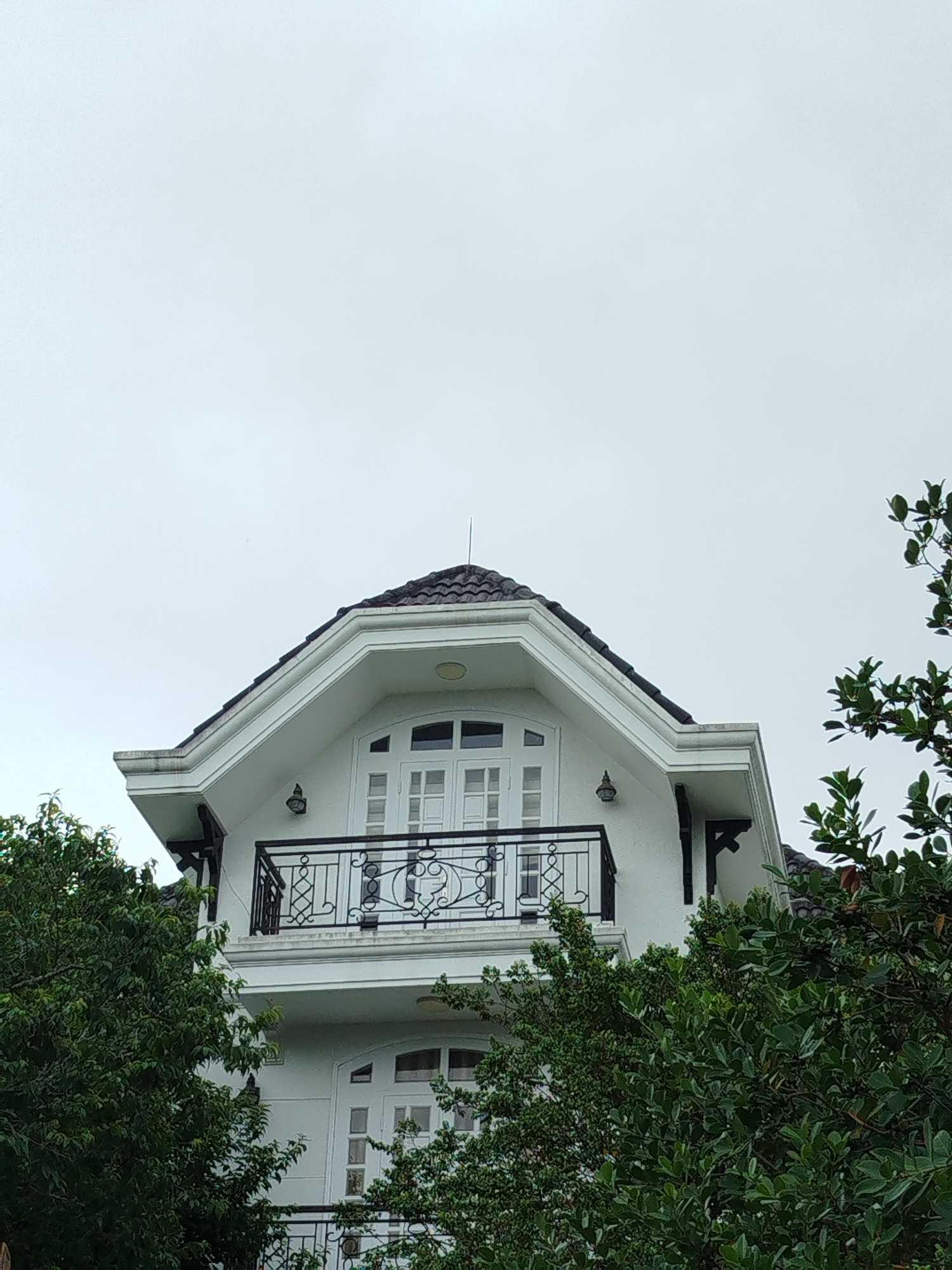 The image depicts a portion of a white, two-story building with a dark gray roof, viewed from below. The building features a balcony on the upper floor with a black metal railing. Windows and doors are white with rectangular panes. Dark supports are visible under the eaves. Green foliage partially obscures the lower portion of the building. The sky is overcast and gray. The architectural style suggests a European influence. The image location is likely in a residential area of Lam Vien - Da Lat, Vietnam based on the architecture and foliage.