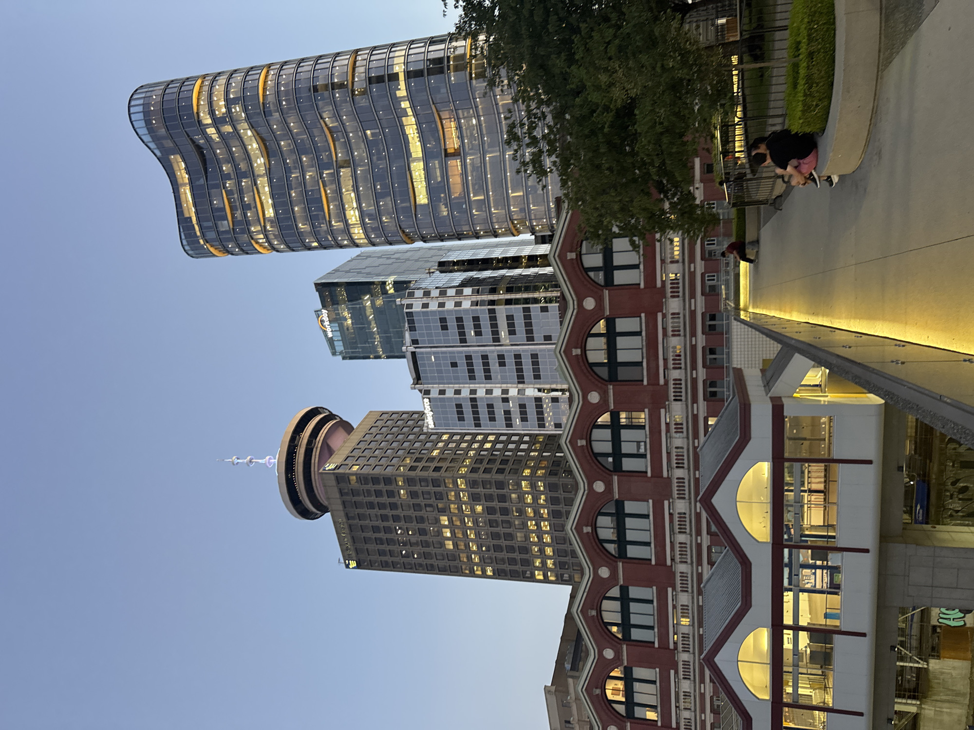 The image depicts a city scene in Vancouver, Canada, at dusk. In the background, several modern skyscrapers rise against a clear, light blue sky. The most prominent building is a tall, square-shaped tower topped with a distinctive circular structure featuring a spire. To its right, a contemporary, curved glass building glows with interior lights. Another, sleeker glass skyscraper with a "Workday" logo is also visible.
In the mid-ground, a red brick building with arched windows and decorative white accents runs horizontally across the frame. Above this, a modern transit station structure with a white facade and large arched windows is illuminated by warm yellow lights.
In the foreground, a paved walkway slopes downwards from right to left, with a concrete barrier along the left edge. Two people, a couple, are seated on a low concrete wall on the right side of the walkway, facing the camera. Another person is sitting alone on a bench further back on the walkway. Green trees and manicured bushes are interspersed throughout the scene, providing a contrast to the urban architecture. The overall impression is one of a bustling urban environment transitioning into evening.