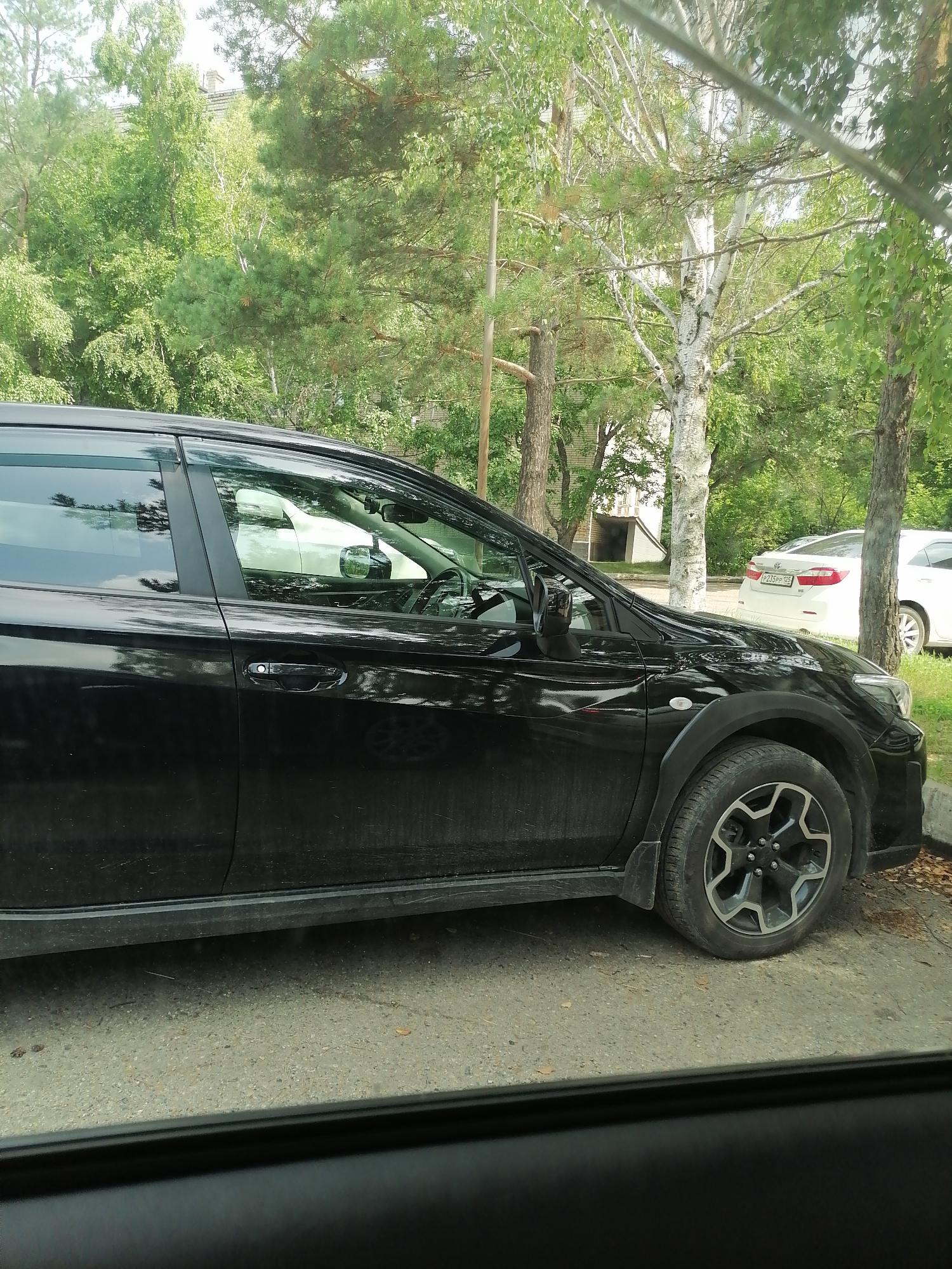 The image shows a black Subaru crossover parked on a paved surface near trees and foliage. A white sedan is visible in the background. The scene appears to be in a residential area, possibly with buildings visible through the trees. The black car is the primary focus, viewed from its side. The environment is natural with an urban element, consistent with a street or parking lot in Lesozavodsk, Russia.