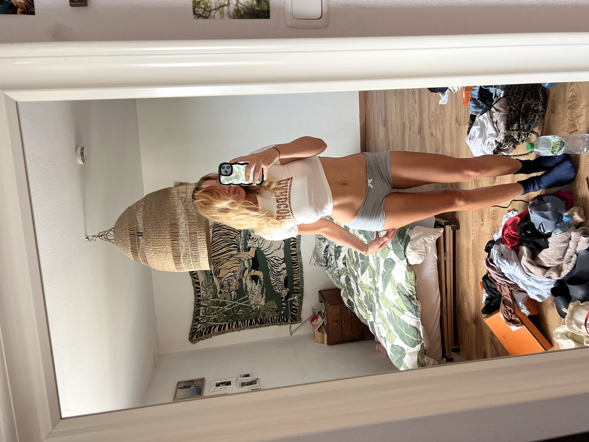 This media file is a mirror selfie taken in a bedroom. The room has wooden floors and a bed with green and white leaf-patterned bedding. A woven basket-shaped lamp hangs from the ceiling. A tapestry depicting tigers hangs on the wall. There are framed photos on the wall and a smoke detector. A pile of clothes is on the floor near the bed, along with a bottle of water and an orange box.
The subject is a young woman with blonde hair, wearing a white crop top and grey shorts. She is holding a smartphone, taking the picture. Her reflection shows her from the waist up, with her legs visible in the lower part of the image. The scene appears to be casual and relaxed, possibly at home. The lighting suggests it might be daytime, though no specific time can be determined. There is no visible text in the image, other than potentially on the smartphone screen. The location context is Mühldorf a. Inn, Germany.