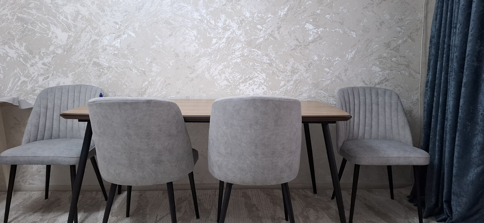 A dining set is positioned in a room. The set includes a light-brown wooden table with dark legs and four light-gray chairs. Two chairs are visible from the front and two from the back. The chairs have a channeled design on the backrest. Behind the dining set, the wall is covered with textured wallpaper featuring a marbled pattern in shades of white and beige. To the right of the dining set, a dark blue velvet curtain hangs. The floor appears to be wood-effect laminate. The image is captured from a slightly low angle, with the table and chairs filling the foreground and midground. The lighting is neutral, suggesting an indoor setting. The scene does not provide any information about the city of Navoiy City, Uzbekistan.