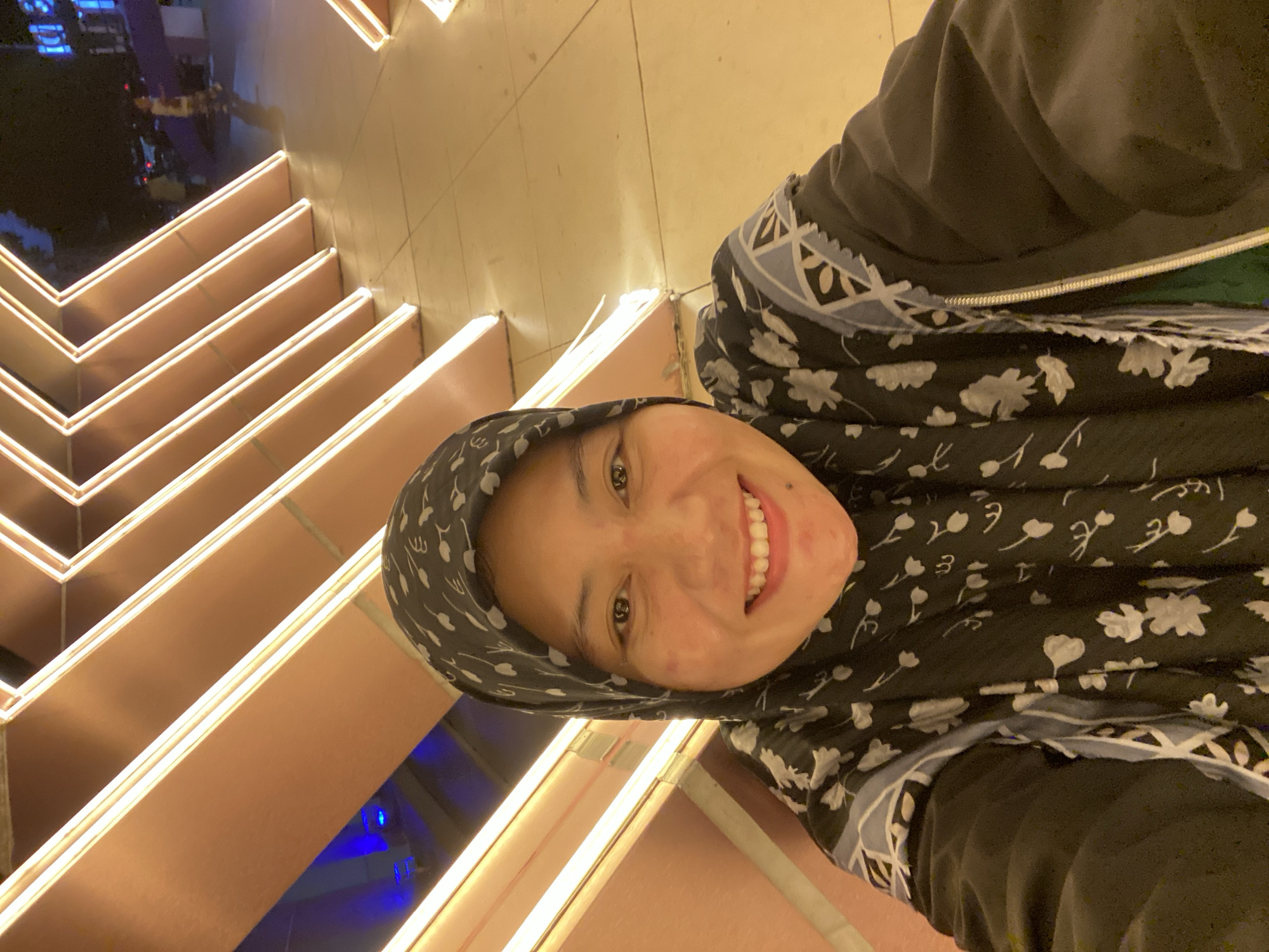 A woman wearing a black hijab with a floral pattern and a black jacket is taking a selfie at night in an outdoor urban setting. The background features a geometric structure with illuminated orange-toned lights and a tiled floor. The woman is smiling at the camera. The scene suggests a public space, possibly a plaza or entertainment area in Banjarbaru, Indonesia, given the stylistic elements and the potential presence of street lights and buildings in the background.