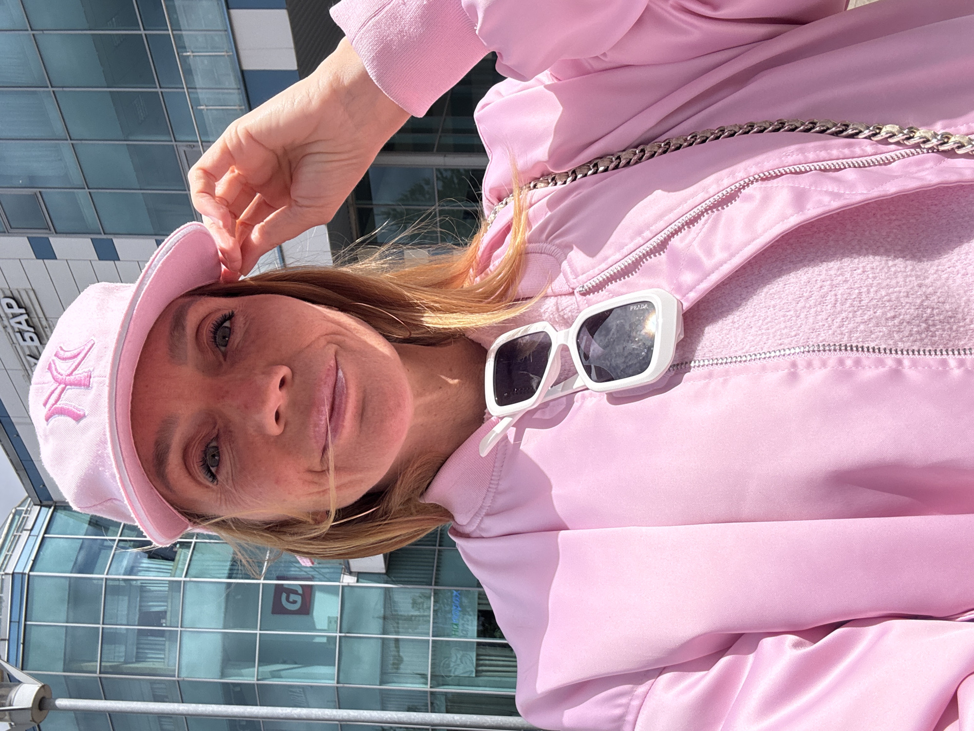 A medium eye-level shot shows a light-skinned blonde woman in a light pink jacket, shirt, and baseball cap, with white Prada sunglasses hanging off the top of the jacket, and a silver chain purse strap over her shoulder. The background is a modern glass-paneled building, reflecting a sunny sky, likely taken outside in Saint Petersburg, Russia.
