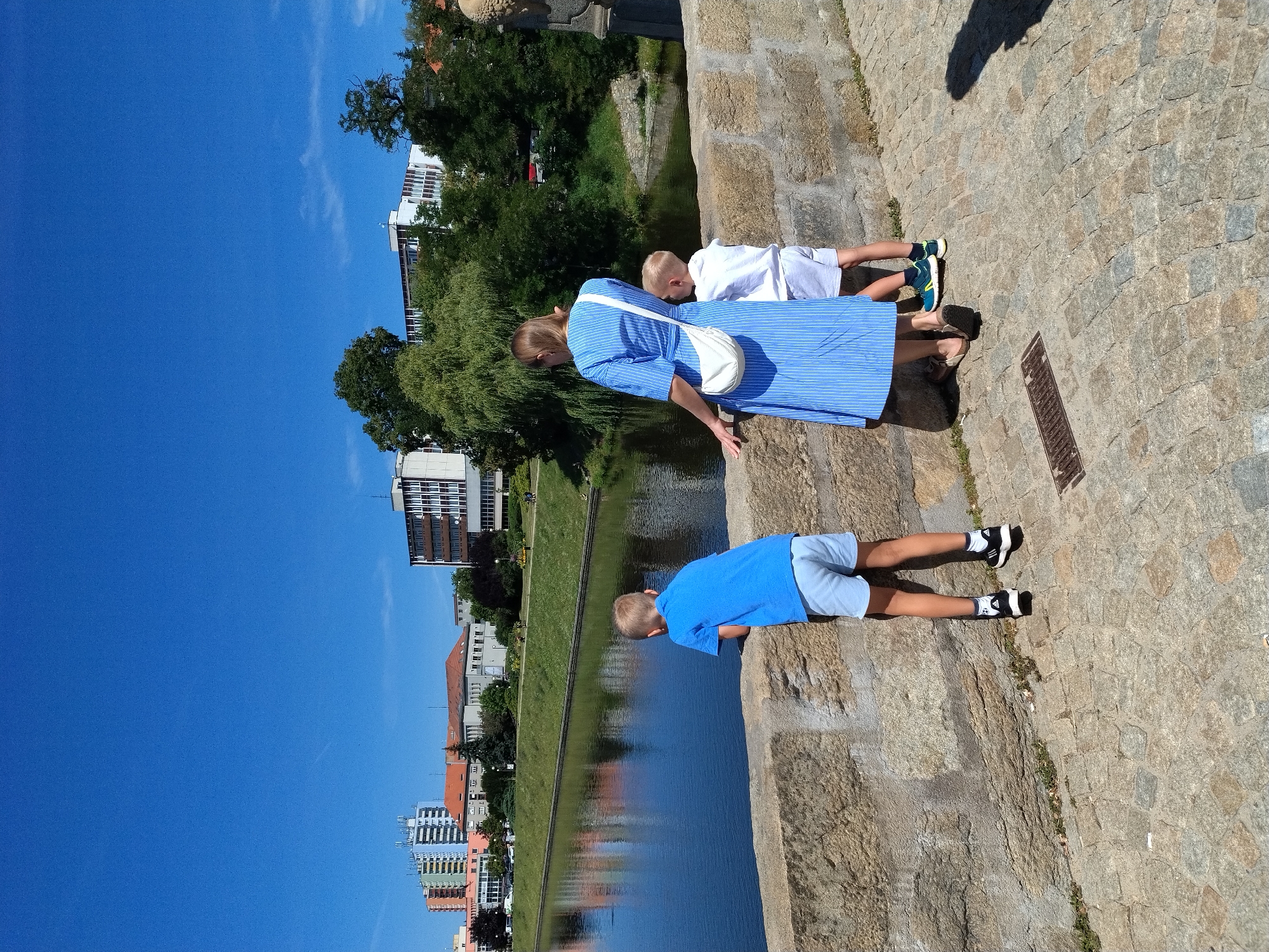 This image depicts a family on a bridge over the Otava River in Písek, Czechia. A woman, wearing a blue and white striped dress and a white crossbody bag, stands with her back to the camera, looking out at the river. To her right, a young boy in a white t-shirt and grey shorts stands next to her, also facing the river. To the woman's left, another boy in a blue t-shirt and light blue shorts leans on the stone bridge railing, gazing into the water. The bridge is constructed of large, rough-hewn stone blocks, and the walkway is paved with cobblestones. In the background, buildings and lush green trees line the opposite bank of the river, with a clear blue sky overhead. The reflection of the buildings and trees is visible on the calm surface of the river.