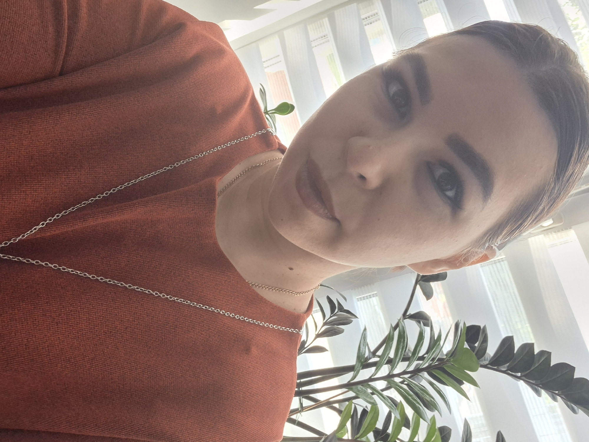 A medium shot taken from a low angle captures a woman wearing a rust-colored sweater and a layered silver chain necklace. She gazes directly at the camera with a neutral expression. In the background, a window is partially obscured by white blinds, revealing glimpses of the exterior, which is in the city of Chaikovsky, Russia. A leafy green plant occupies the lower right corner of the frame.