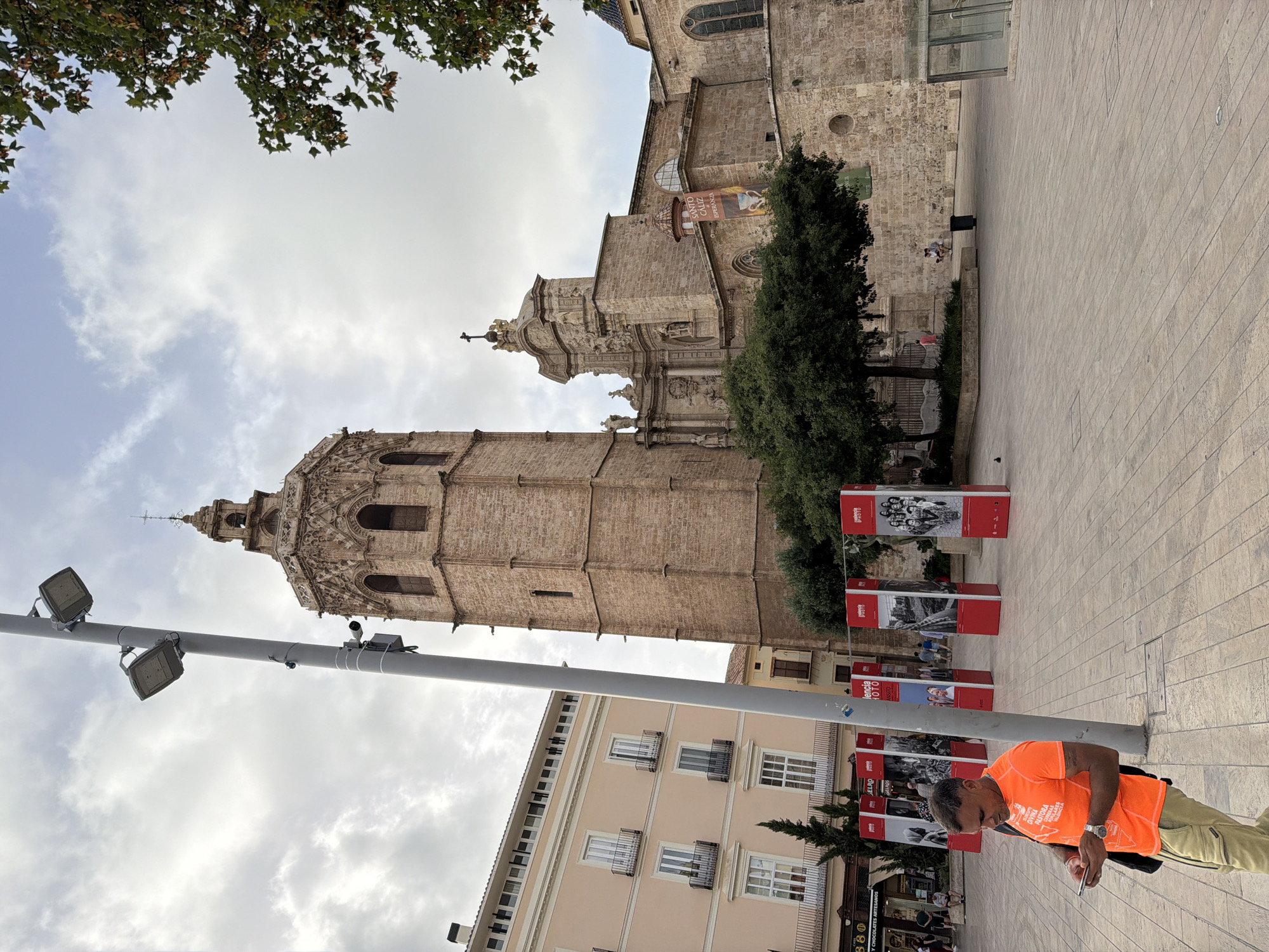 A man in an orange t-shirt and khaki pants is looking at his phone while standing on a paved square in front of a large stone building with a tall tower. The building appears to be a cathedral or church. There are several tall red rectangular displays with black and white photos on them in the square. A large green tree is in front of the building, partially obscuring it. The sky is cloudy.