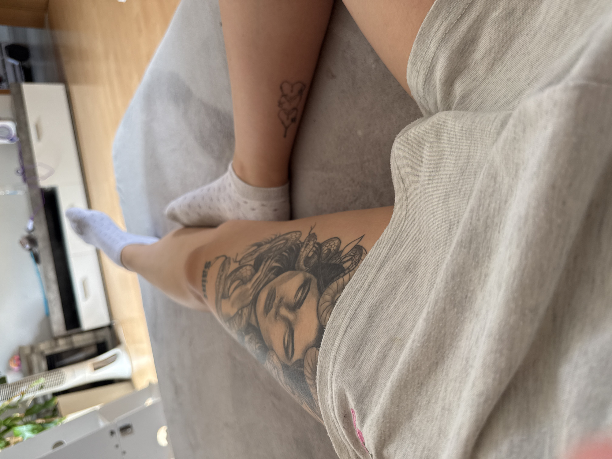 The image shows a person's legs resting on a grey couch. The person is wearing light-colored socks and grey shorts. There are tattoos visible on both legs. One tattoo on the left thigh is a portrait of a face surrounded by snakes, which appears to be Medusa. A smaller tattoo of three cloud-like shapes is visible on the right calf. In the background, a white fan and a modern entertainment center with a wooden floor can be seen. The lighting suggests the photo was taken indoors during the day.