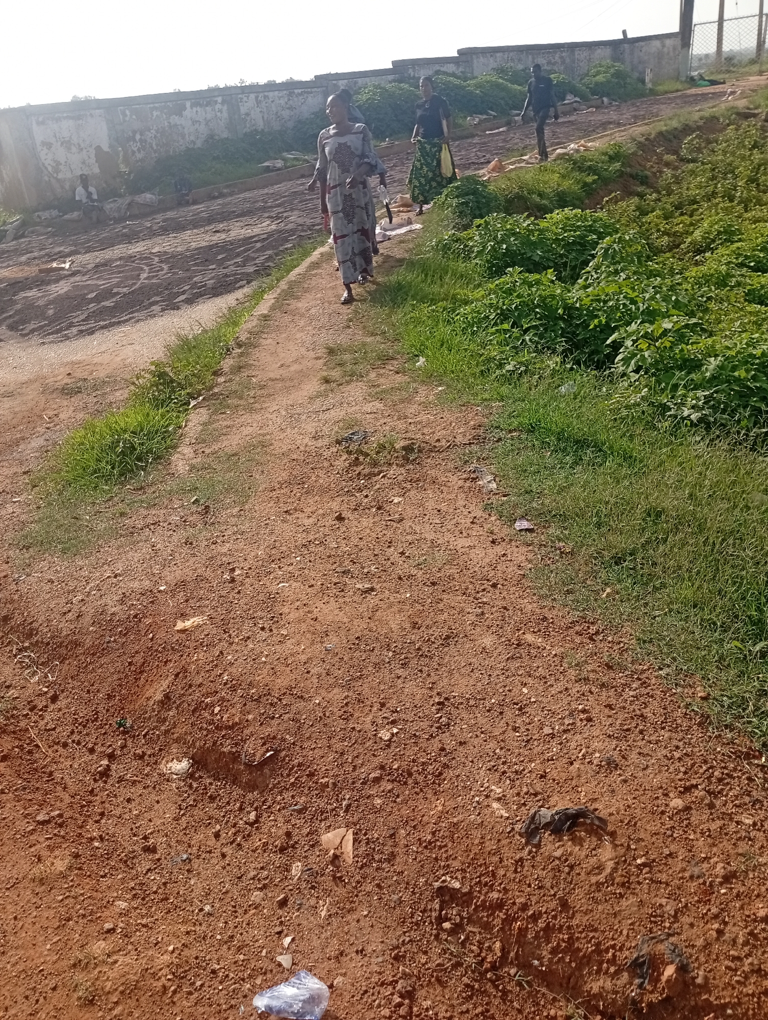The image shows an outdoor scene on a dirt path, likely in Rafin Par, Nigeria, with a concrete wall in the background. In the middle ground, three people are visible walking away from the camera. The person in the foreground is a woman wearing a long, patterned dress. Behind her, another woman in a patterned skirt and dark top walks alongside a man in a dark shirt and pants. To the left of the path, near the concrete wall, a person is seated, and further left, there are some discarded items and debris. The path is uneven and covered in reddish-brown dirt, with patches of green grass growing along the edges. The background features the concrete wall, some vegetation, and a bright, overcast sky.