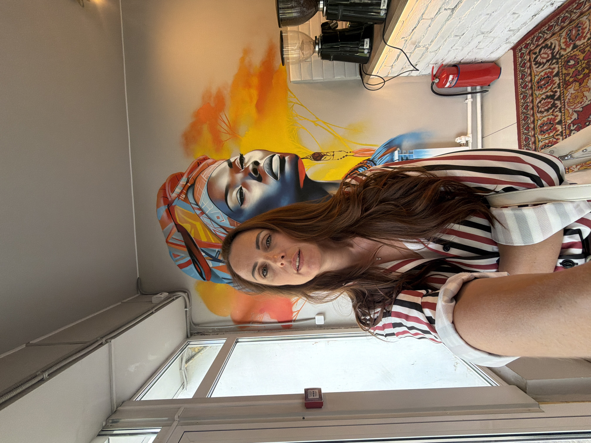 A medium shot captures a woman with long brown hair taking a selfie in front of a vibrant mural of a woman's face wearing a colorful headwrap. She is wearing a striped shirt with red, black, and white stripes and a white cuff. The mural is on a gray wall, with orange and yellow abstract shapes and yellow tree-like figures in the background. To the right of the mural are two black coffee grinders on a wooden counter, with white brickwork behind them. A red fire extinguisher is visible on the floor to the right, next to a rug with an intricate red, black, and yellow pattern. The woman is standing near a window with a white frame, and a red fire alarm is mounted on the left side of the frame. Electrical conduits are visible along the ceiling and walls. The scene appears to be indoors, possibly in a cafe or shop. The location is not specified in the image or provided hints.