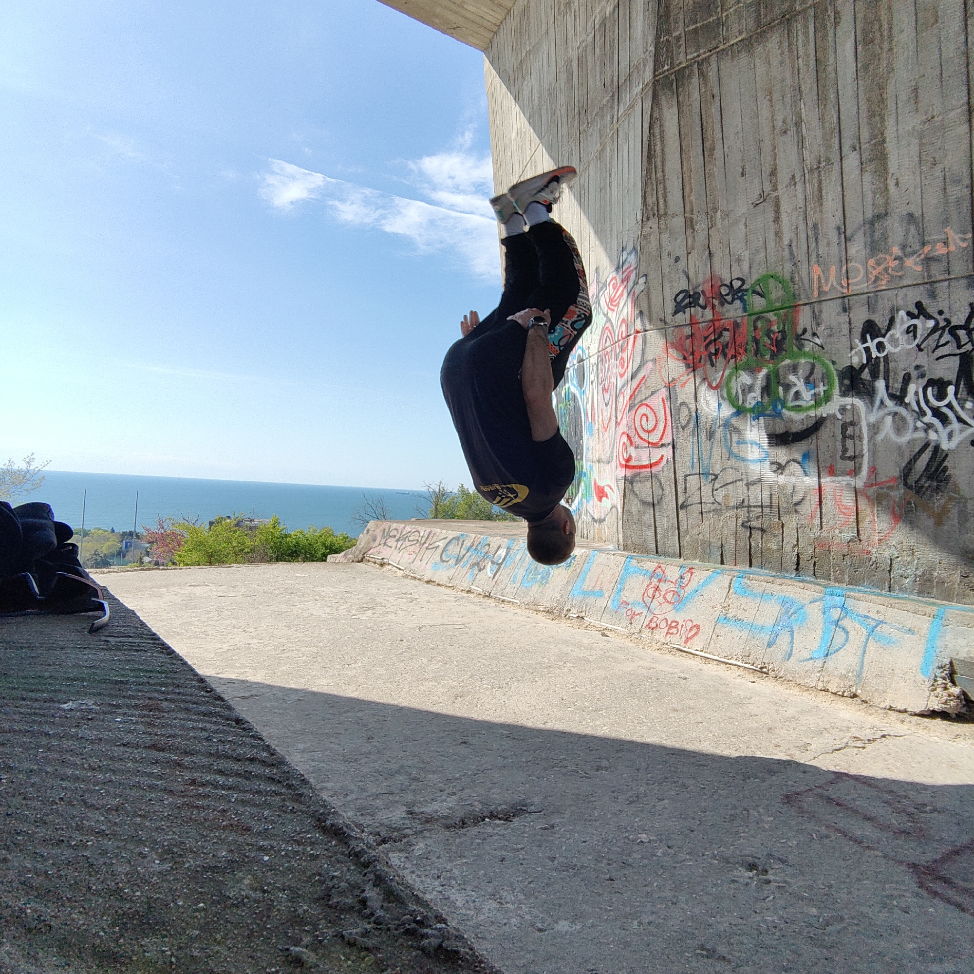 A man performs a backflip in Varna, Bulgaria, under a concrete structure with graffiti. The sky is blue with light clouds. The sea and some green foliage are visible in the background. The man is wearing dark pants and a black t-shirt with a design on it. His sneakers are visible above him as he flips, and the ground below is concrete.