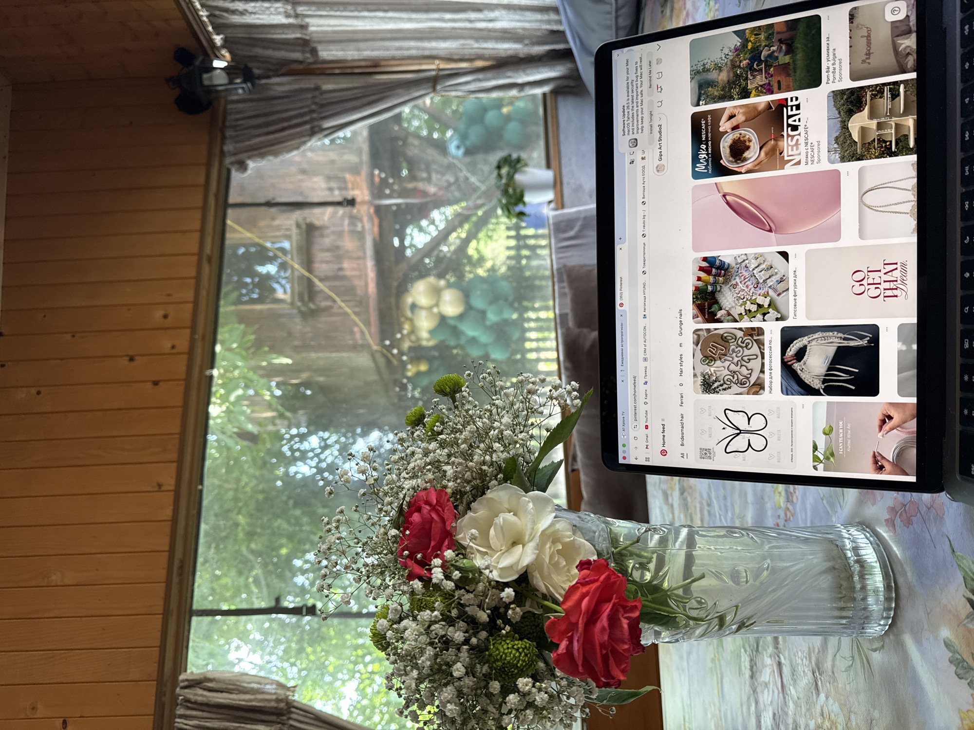The image captures a serene indoor scene with a view to the outdoors, illuminated by natural daylight. The setting appears to be a cozy room, possibly a cabin or a home, characterized by light brown wooden paneled walls. A large window dominates the left side, offering a glimpse of lush green foliage and trees outside, with what looks like balloons or round decorations suspended in the distance, and a rustic wooden structure, possibly a treehouse or shed, visible through the window.
In the foreground, a vibrant bouquet of flowers sits in a clear, fluted glass vase on a light-colored, patterned tablecloth. The bouquet features several red and white roses, interspersed with numerous delicate white baby's breath (Gypsophila) flowers and clusters of green chrysanthemum-like blooms.
To the right, a black laptop (MacBook) is open, displaying a Pinterest feed. The screen shows a variety of images, including a NESCAFÉ advertisement with text "Мляко с NESCAFE®" (Milk with NESCAFE), a "GO GET THAT Dream." graphic, pictures of craft items like knitted art and plaster figures, and various fashion and beauty-related content. A "Software Update" notification for "macOS Tahoe 14.5" is also visible at the top of the screen, with options to "Install Tonight" or "Remind Me Later." Other visible text on the browser tabs and search bar includes "A1 Xplore TV," "Ежедневни астропрогнози" (Daily horoscopes), "Pinterest," "Автокъща HYUND." (Hyundai car dealership), "Витоша Ауто ЕООД" (Vitosha Auto EOOD), "Карти" (Maps), and "Превод" (Translate), indicating a user engaging with online content in Bulgarian and English. The overall atmosphere suggests a moment of relaxation or casual work within a pleasant domestic environment.