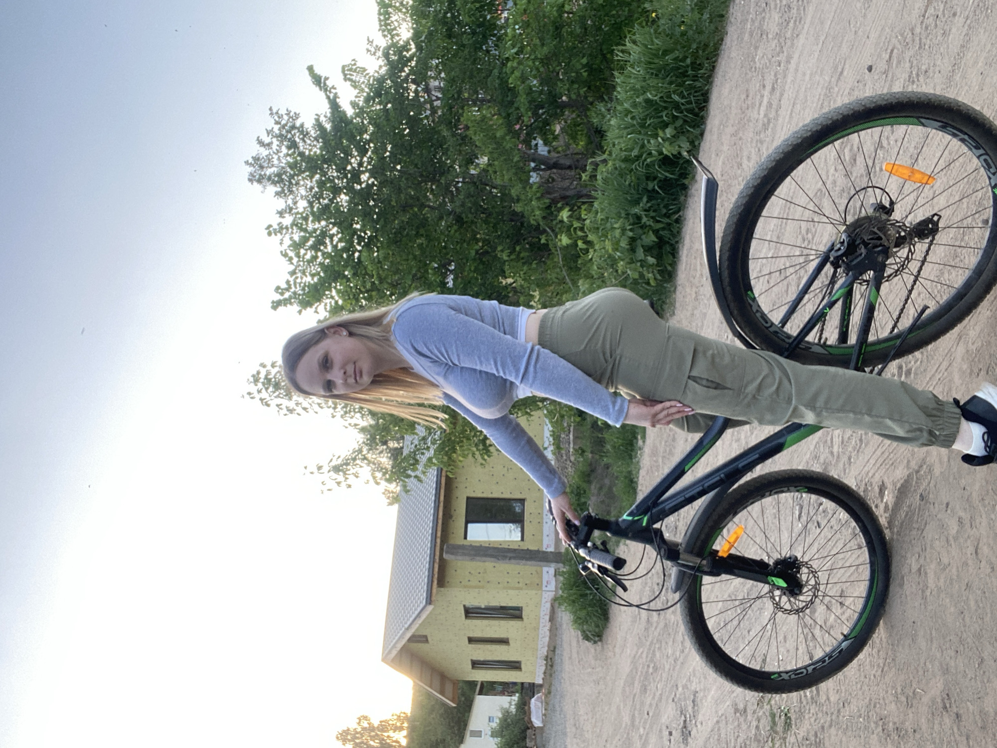 A woman is standing next to a bicycle on a dirt path. The woman is wearing a light grey long-sleeved shirt and green pants. She has long blonde hair. The bicycle is black and green. In the background is a building under construction with yellow siding and a few trees. The sky is light and overcast. The location is Chaikovsky, Russia.