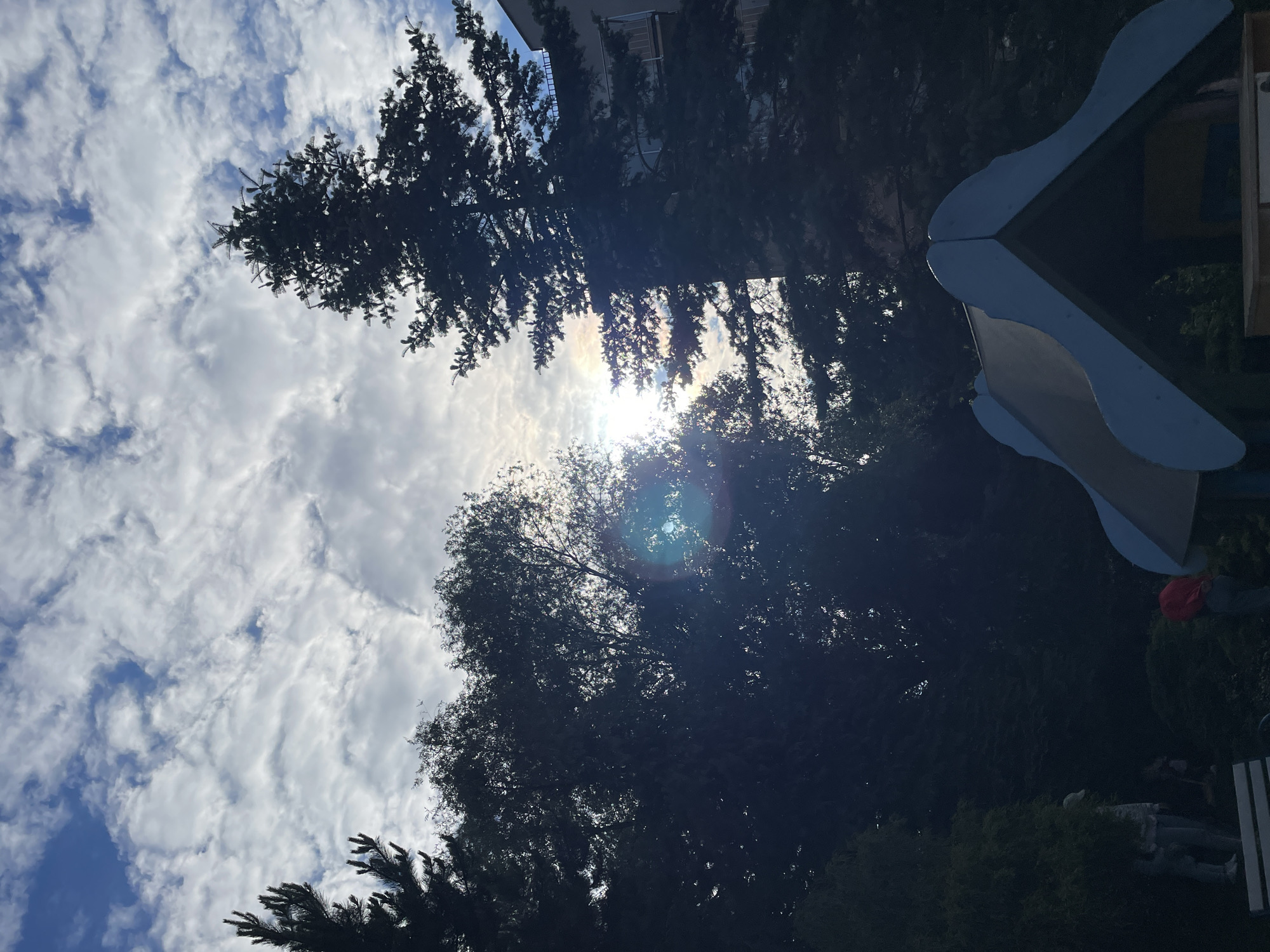 An outdoor, low-angle shot captures a sky filled with a mix of bright white clouds and patches of blue, with the sun partially obscured by trees, causing a lens flare. Silhouetted against the sky are the dark green branches of coniferous trees on the left and right, and leafy trees in the middle. In the foreground, partially visible, is the roof of a playground structure, painted in shades of blue and grey. To the left, on a bench, are two individuals, one wearing a light-colored baseball cap and a patterned shirt, and the other in a light-colored hoodie. To the right, a person in a red jacket is visible near the playground structure. In the background, a multi-story building is visible behind the trees on the right. The overall lighting suggests it is daytime.