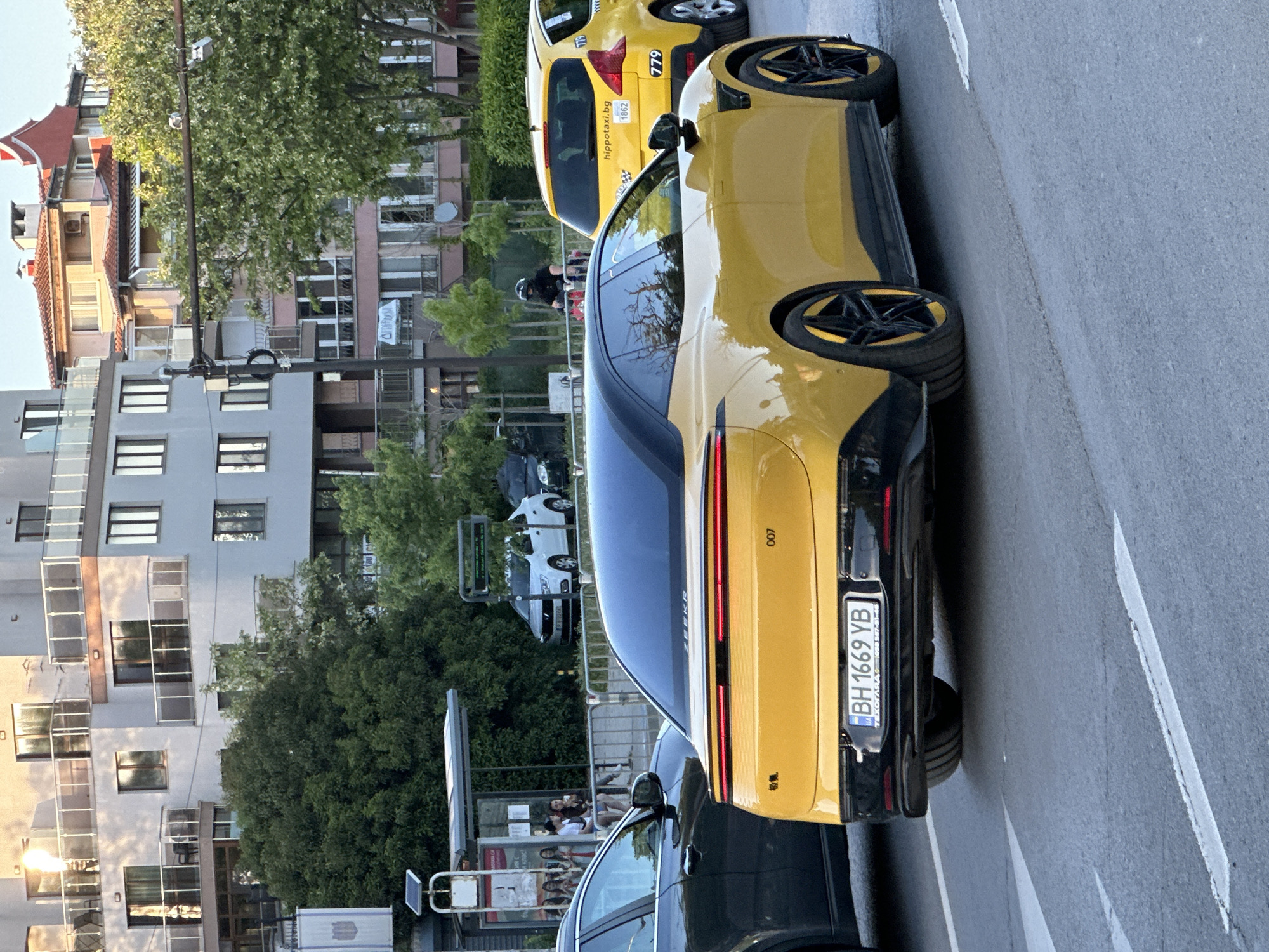 The image depicts a street scene in Varna, Bulgaria. A yellow Porsche Taycan with license plate "BH 1669 YB" is parked on the right side of the road. A yellow taxi is visible behind the Porsche. People are waiting at a bus stop on the left side of the road. Buildings and trees form the backdrop of the scene. The photo is taken from a low angle.