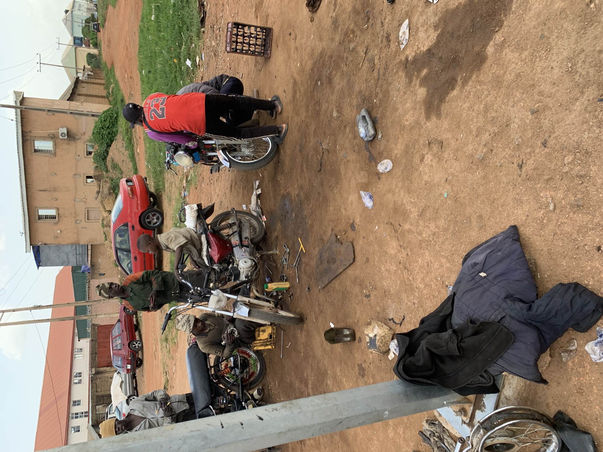 The image captures an outdoor scene in Jos, Nigeria, likely a roadside workshop or informal gathering. The ground is dirt, with some sparse grass and scattered debris, suggesting an unpaved area. The lighting and shadows indicate it's daytime, possibly late morning or afternoon, with clear weather.
Several individuals are present, engaged in what appears to be motorcycle repair or maintenance. One person in a red shirt with "23" on the back is working on a motorcycle, while others are scattered around, some looking on, others also working. There are at least three motorcycles visible in various states of repair, along with tools and parts laid out on the ground. A red car is parked nearby, and in the background, buildings and a power line can be seen, providing context of a semi-urban or rural environment.
One of the notable details is a dark blue jacket and a black garment laid out on the ground, possibly belonging to one of the individuals or left there for a moment. A crate filled with what look like small items, possibly nuts or bolts, is also visible. The overall atmosphere seems focused and practical, with the individuals appearing concentrated on their tasks. There is no visible text in the image.
