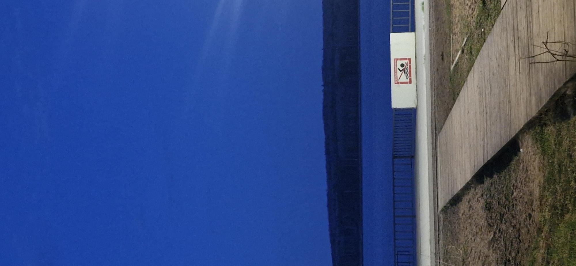 The image shows a vertical shot of what appears to be a portion of a waterfront area. On the left is a large expanse of blue, suggesting water, possibly a river or lake. Moving rightward, a structure of dark gray or black, perhaps a wall or embankment, is visible, followed by a light-colored section with a safety sign that depicts a figure being swept away by water. A blue ladder is attached to the structure. Next to that is a wooden walkway or path, and adjacent to the walkway is an area of grass and vegetation. The location is Unknown, Unknown.