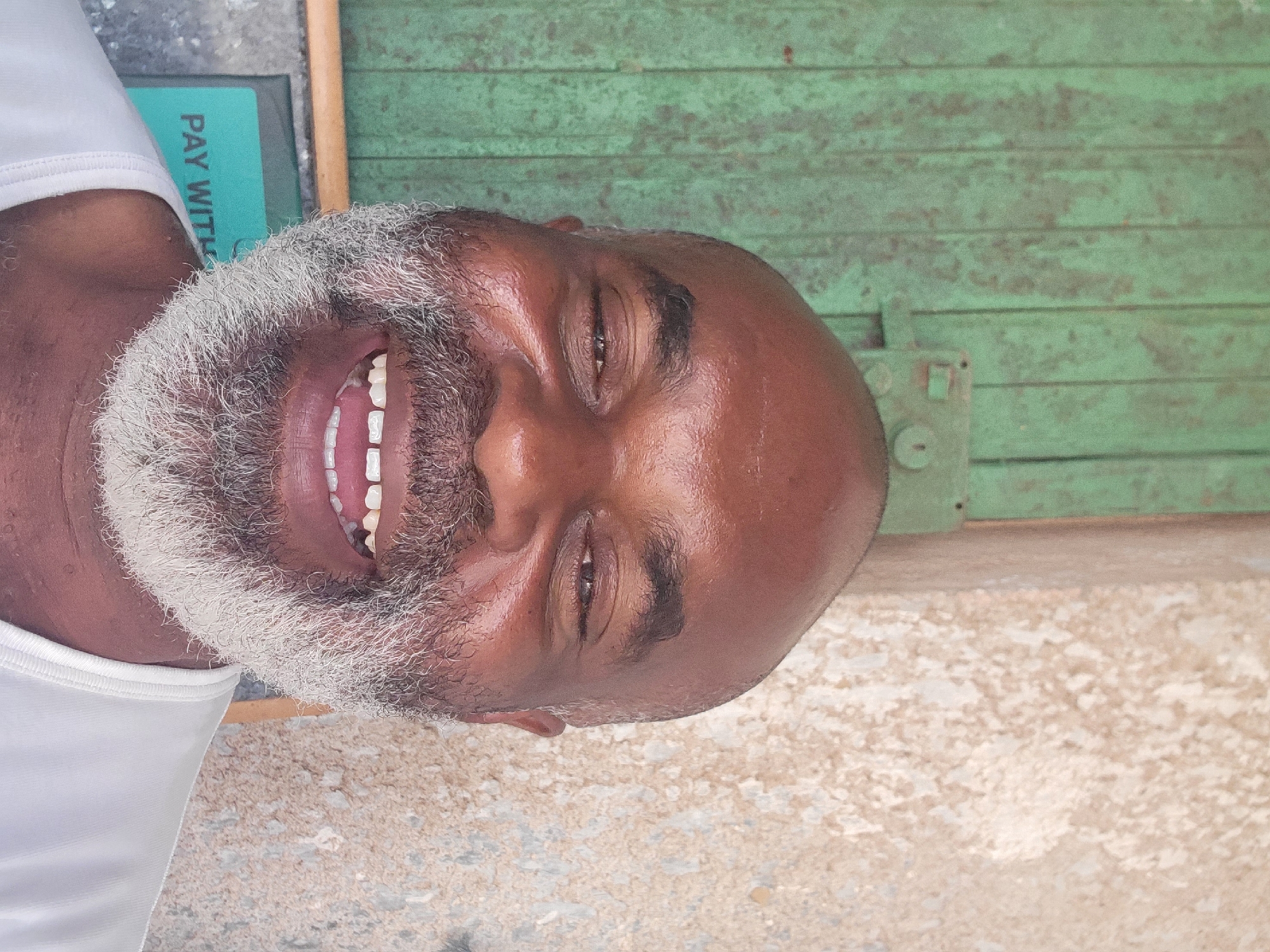 This portrait captures a smiling man in Maiburiji, Nigeria, seen from the chest up. He has dark skin, a bald head, and a full, distinctive white and grey beard. He is wearing a white sleeveless top, partially visible on his left shoulder. His eyes are squinted, and his skin appears shiny, possibly from sweat or heat, as he beams with a wide smile, revealing his teeth.
The background behind him features a weathered, light green wooden surface, likely a door or wall, with visible horizontal planks and a metal latch mechanism on the right side. To the upper left of his head, a blue-green rectangular object is partially visible, on which the white text "PAY WITH" can be read. The lower right portion of the image shows a light-colored, textured ground, suggesting an outdoor setting. The bright lighting indicates it is daytime, and the man's broad smile conveys a joyful or amused emotion.