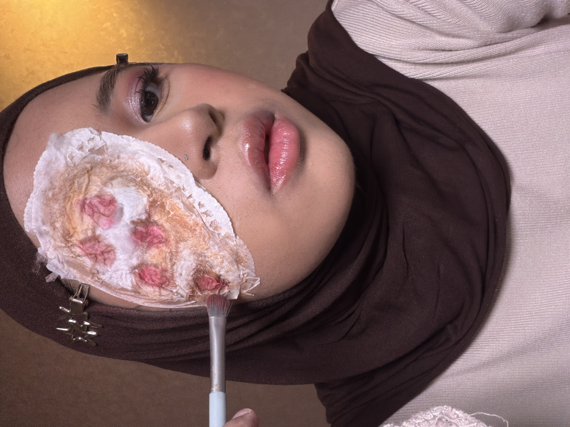 A young woman wearing a brown hijab and a light beige top is lying on her back. She is undergoing makeup application. A wound-like prosthetic is adhered to her forehead and the right side of her face, decorated with reddish-pink spots and tan coloration. A makeup brush is being held in front of the prosthetic. A metal hair clip is visible near her right temple, and a small piercing is present near her left nostril. The background appears to be a textured wall with a golden-brown hue. The image likely originated from East Jakarta, Indonesia.