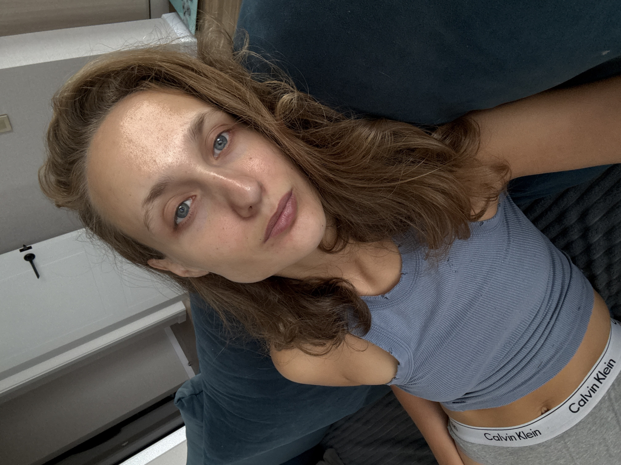A young woman with light brown hair and blue eyes is lying on a dark blue couch. She is wearing a light blue ribbed tank top and grey Calvin Klein underwear. Her right arm is resting on the couch, and her head is propped on her arm. The woman is looking directly at the camera. The background shows a white door with a black handle and a wall with some molding. The image appears to have been taken indoors, likely in a residential setting in the village of Podshivalovo, Russia.