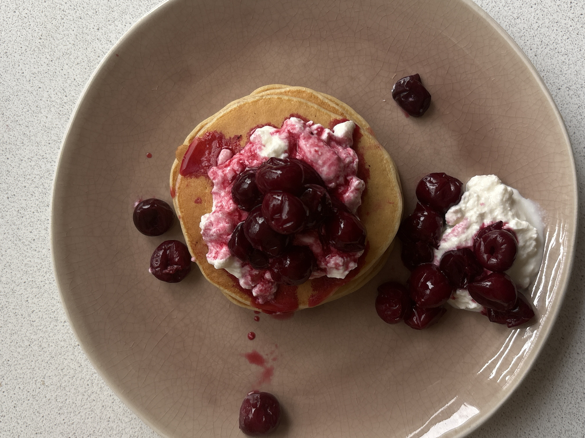 A top-down shot features a plate of pancakes topped with ricotta cheese and cherries. The pancakes are stacked and located in the center of the plate. A portion of ricotta cheese and cherries is placed to the right of the pancakes. Some cherries are scattered around the plate. The plate is light brown with a cracked pattern. The surface under the plate appears to be a white, speckled material. The scene does not contain information to ascertain its location.