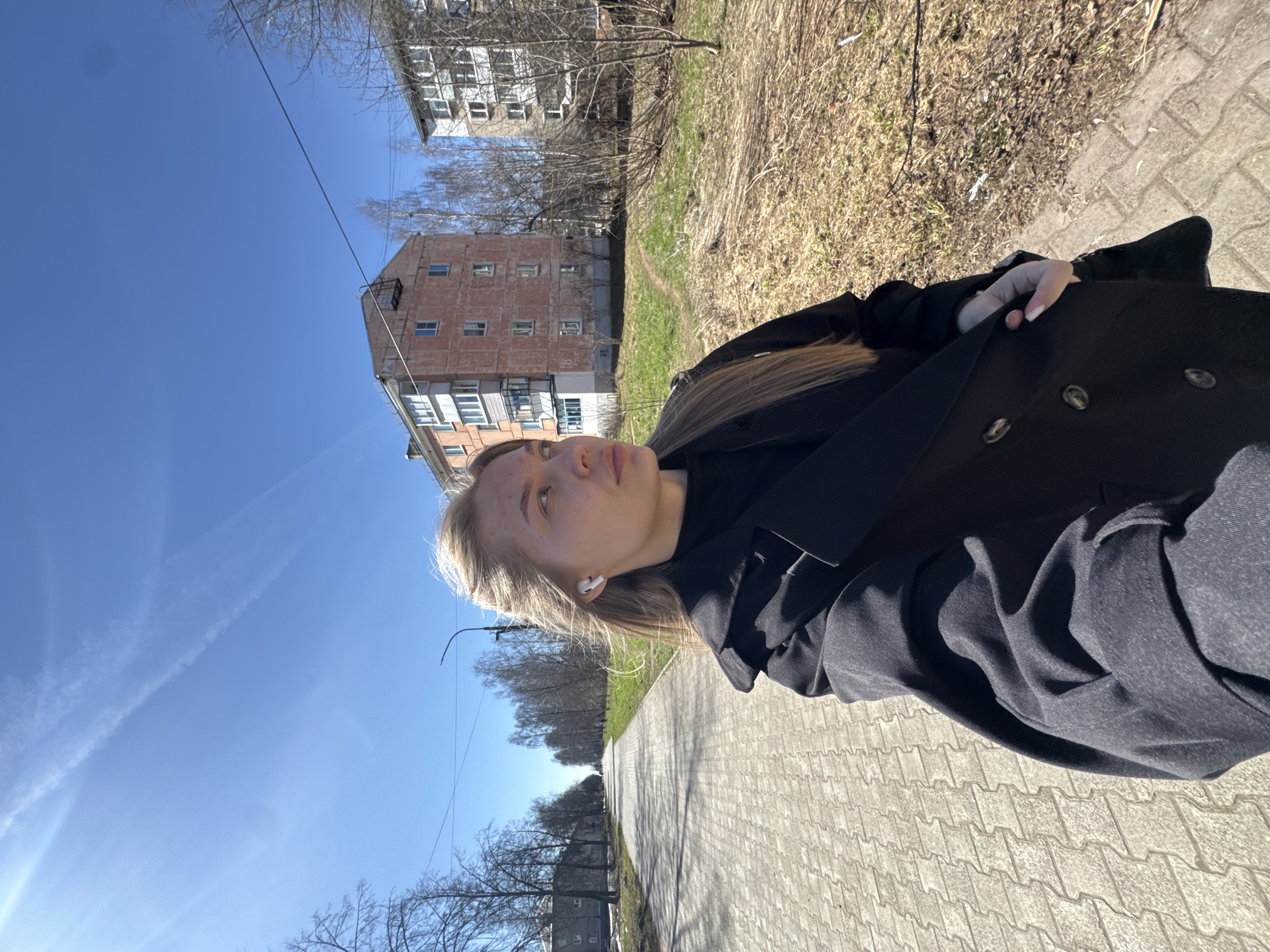 A young woman with blonde hair is standing on a paved sidewalk in Chaikovsky, Russia. She is wearing a black coat and has a white wireless earbud in her ear. The background includes buildings, trees, grass, and a blue sky with contrails. The camera angle is low, possibly a selfie. The setting appears to be an urban environment, possibly a residential area.