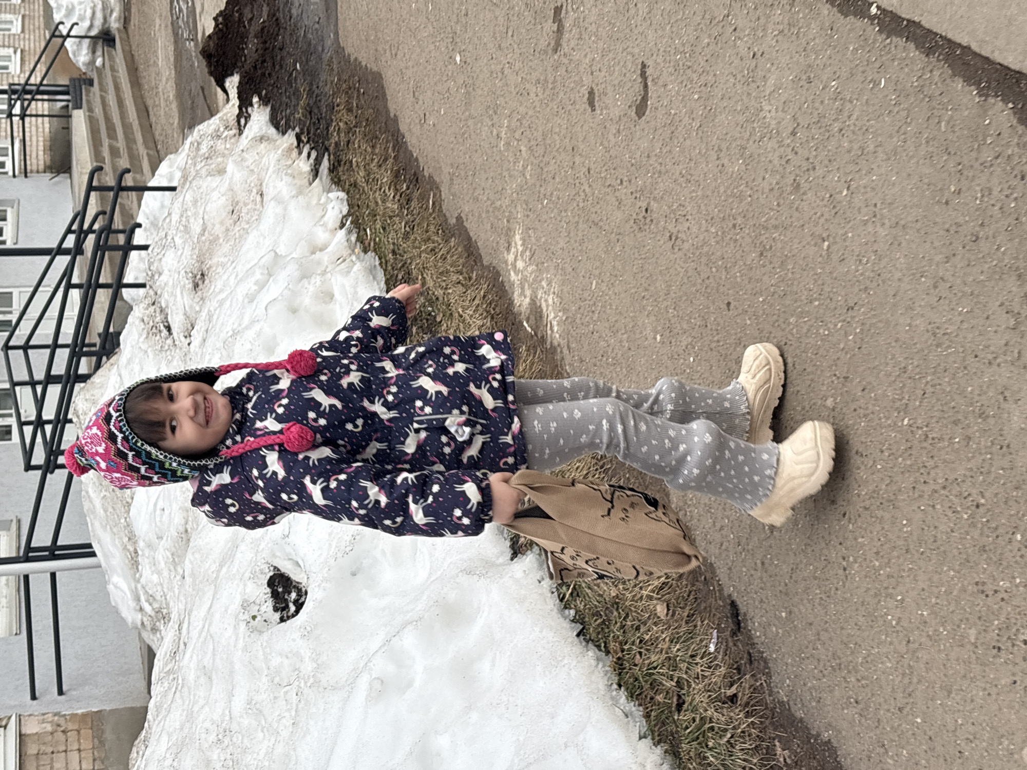 An image taken in the city of Chaikovsky, Russia, depicts a young child walking outdoors during what appears to be late winter or early spring. The child, approximately 4-6 years old, is positioned slightly right of center, looking directly at the camera with a smile. They wear a multi-colored knit hat featuring pink, white, and dark blue patterns, with ear flaps and two prominent pink pom-poms hanging down. A dark blue winter jacket adorned with white unicorn patterns and small pink accents covers a pair of light grey pants patterned with small white paw prints. The child wears light beige, chunky-soled footwear. In their left hand, they carry a light brown or beige canvas-like tote bag, possibly decorated with dark illustrations.
To the child's left and extending behind them is a large, irregularly shaped mound of melting snow, mixed with dirt and brown foliage. Patches of dry, brown grass are visible between the snow and the paved surface. The ground beneath the child and extending into the foreground is dark grey asphalt or concrete, appearing somewhat damp. In the background, beyond the snow, a light-colored building with multiple windows is visible. A set of concrete stairs with dark metal handrails leads up to an entrance of this building. Another snow accumulation is present to the right of the stairs. The sky is not visible, but the overall lighting suggests an overcast day.