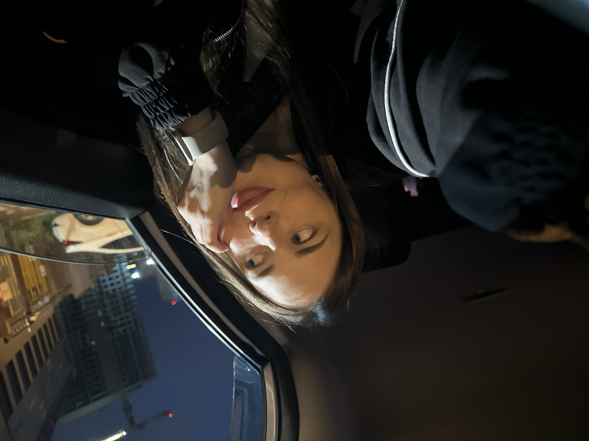 The image shows a young woman sitting in the passenger seat of a car at night, looking out the window. She is wearing a black hoodie with white trim and a white smartwatch. Her face is illuminated by the ambient light from outside, and she has her hand on her chin, with her tongue slightly sticking out. In the background, city lights and buildings are visible, including a crane on a construction site, indicating an urban environment. The interior of the car is dark, with only a dim light visible on the ceiling. The overall impression is a candid shot of a person contemplating while traveling at night in a city.