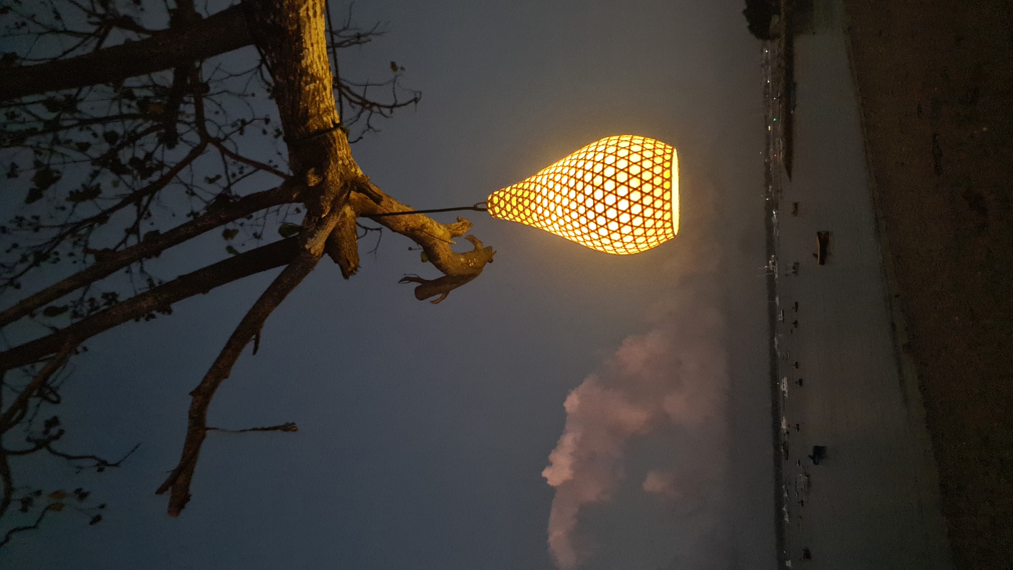 A lamp made of woven material with a crisscross pattern hangs from a tree branch over a beach. The lamp is lit with a warm yellow light. In the background, a body of water is filled with many boats of various sizes. The sky is dark blue with some pinkish clouds on the left side. This scene is likely located at a beach in Denpasar, Indonesia, where such decorative lamps are often used to create ambiance.