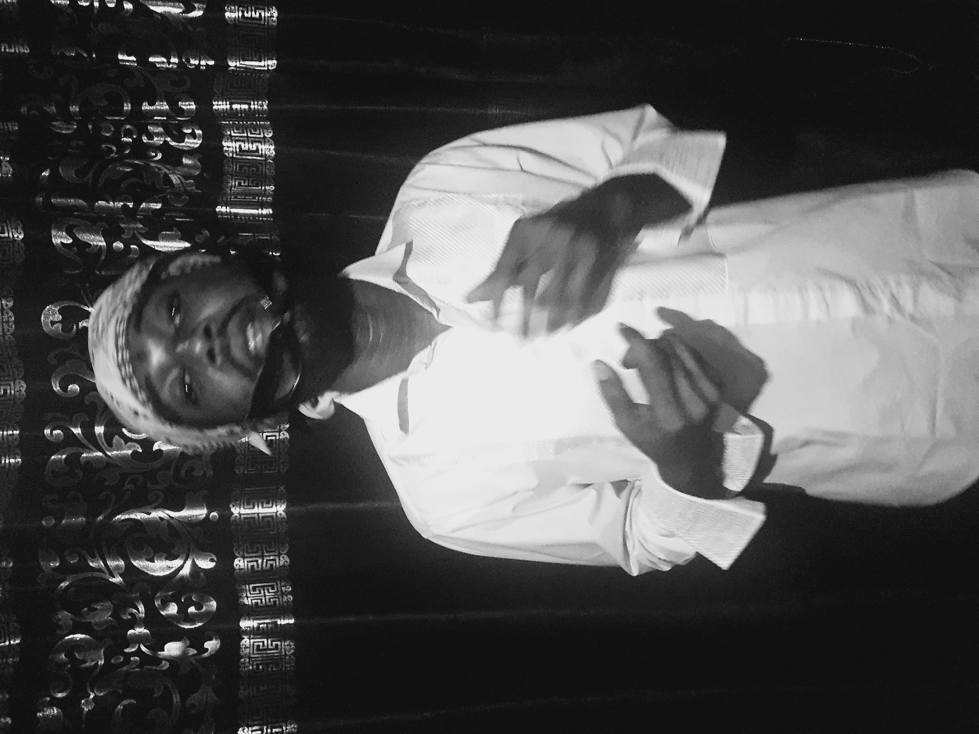 A black and white medium shot depicts a dark-skinned man in a light-colored collared shirt and patterned head covering. He is positioned at an angle, appearing to face the viewer with an upward gaze. His hands are raised, and fingers appear to be interlocked. The background features a dark, patterned fabric. The location, Lokoja, Nigeria, is not discernible from the image content.