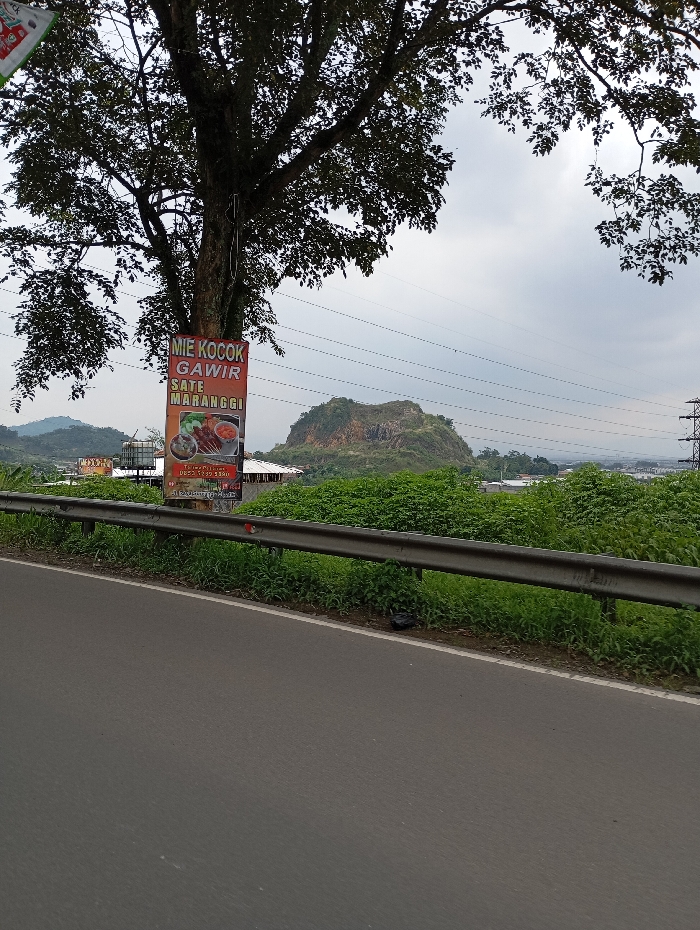 The image shows a roadside view of a paved road in Kutawaringin, Indonesia. A large tree occupies the left side of the image, partially obscuring a sign for a restaurant serving "Mie Kocok Gawir Sate Maranggi." The sign is mounted on a pole and has a red background with white lettering. The road is bordered by a metal guardrail and a grassy verge. In the distance, a rocky hill covered in green foliage rises up on the right side. A power line runs across the top of the image. The sky is overcast. The coordinates provided suggest the location is on Jalan Raya Sampit - Pangkalan Bun, a main road running through Kutawaringin.