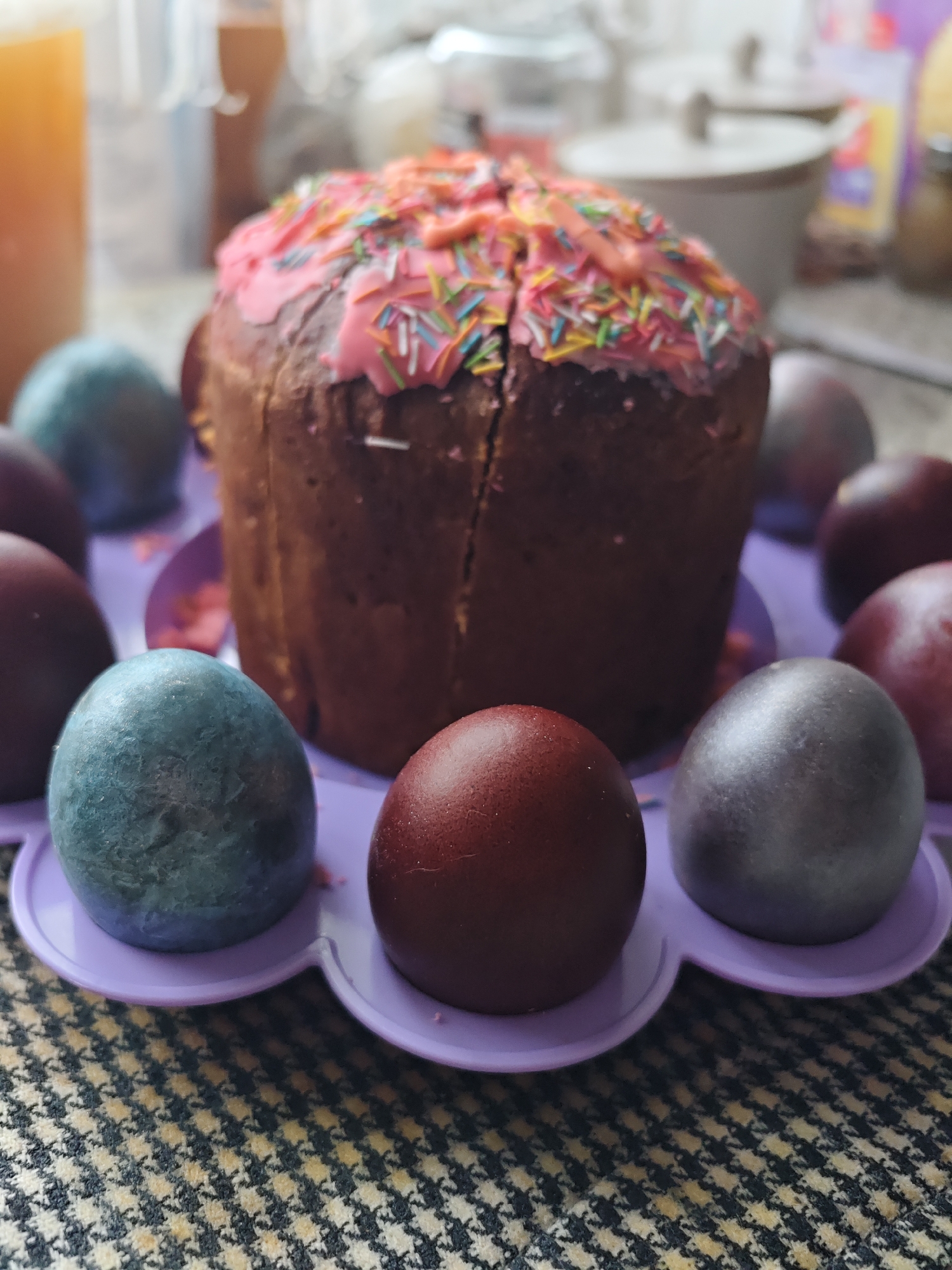 The image shows an Easter cake with pink frosting and colorful sprinkles, surrounded by dyed Easter eggs in a purple plastic egg holder. The eggs are various shades of blue, red, and silver. The scene is set on a surface with a houndstooth pattern. The location might be Уральское, Russia.