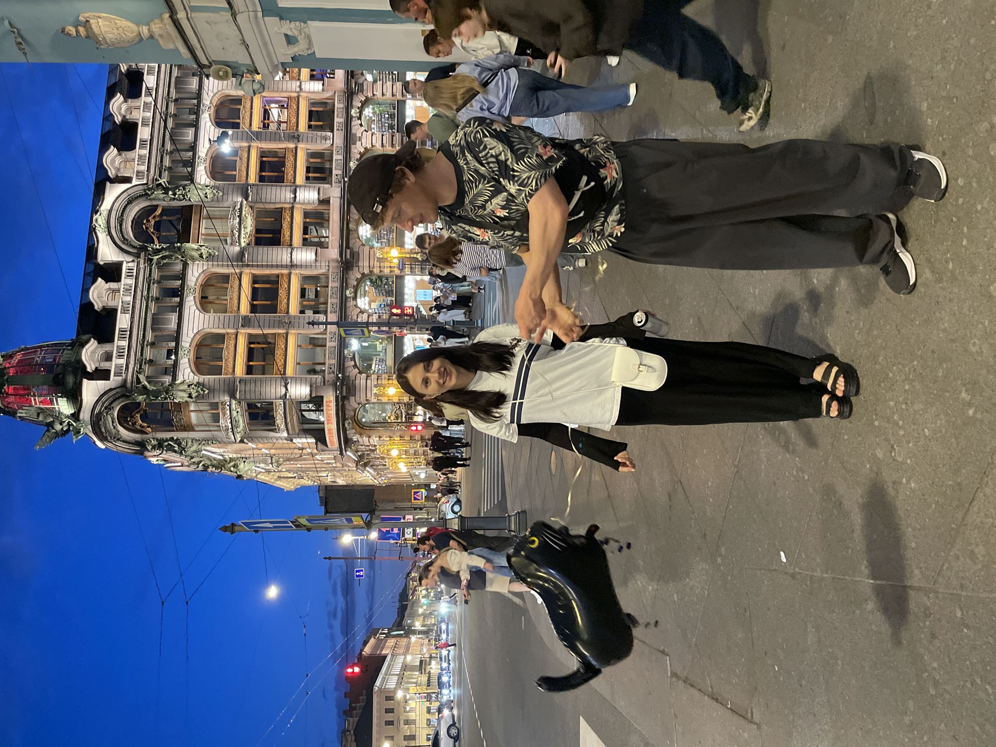 The image depicts an urban street scene at dusk or early evening in Saint Petersburg, Russia, specifically outside the Singer House (Dom Knigi) on Nevsky Prospekt, identifiable by its distinctive architecture and the visible "КОМПАНІЯ ЗИНГЕРЪ" inscription. The sky is a deep blue, and various buildings and streetlights are illuminated.
In the foreground, a woman stands on the left, smiling towards the camera. She wears a white t-shirt with dark stripes over a black long-sleeved shirt, black wide-leg pants, and black platform sandals. A white crossbody bag is slung over her shoulder. She holds the string of a large, black, cat-shaped helium balloon with yellow eyes, which floats slightly above the sidewalk between her and the man next to her.
To her right, a man faces the woman, gesturing with both hands, suggesting an interaction. He wears a dark baseball cap, a dark t-shirt with a tropical floral pattern, dark pants, and dark athletic shoes with white soles. A black fanny pack is visible across his torso.
The background is populated with numerous pedestrians on the sidewalk and a street with vehicles. Several streetlights are active, casting light onto the scene. Red traffic lights are visible further down the street. Overhead wires crisscross the sky. The Singer House, an ornate multi-story building, dominates the right background, displaying illuminated windows and intricate architectural details. On the far right, a segment of a light-colored building with a classical urn decoration is visible. The ground is paved with large, light-colored slabs, and shadows are cast by the people and the balloon.
