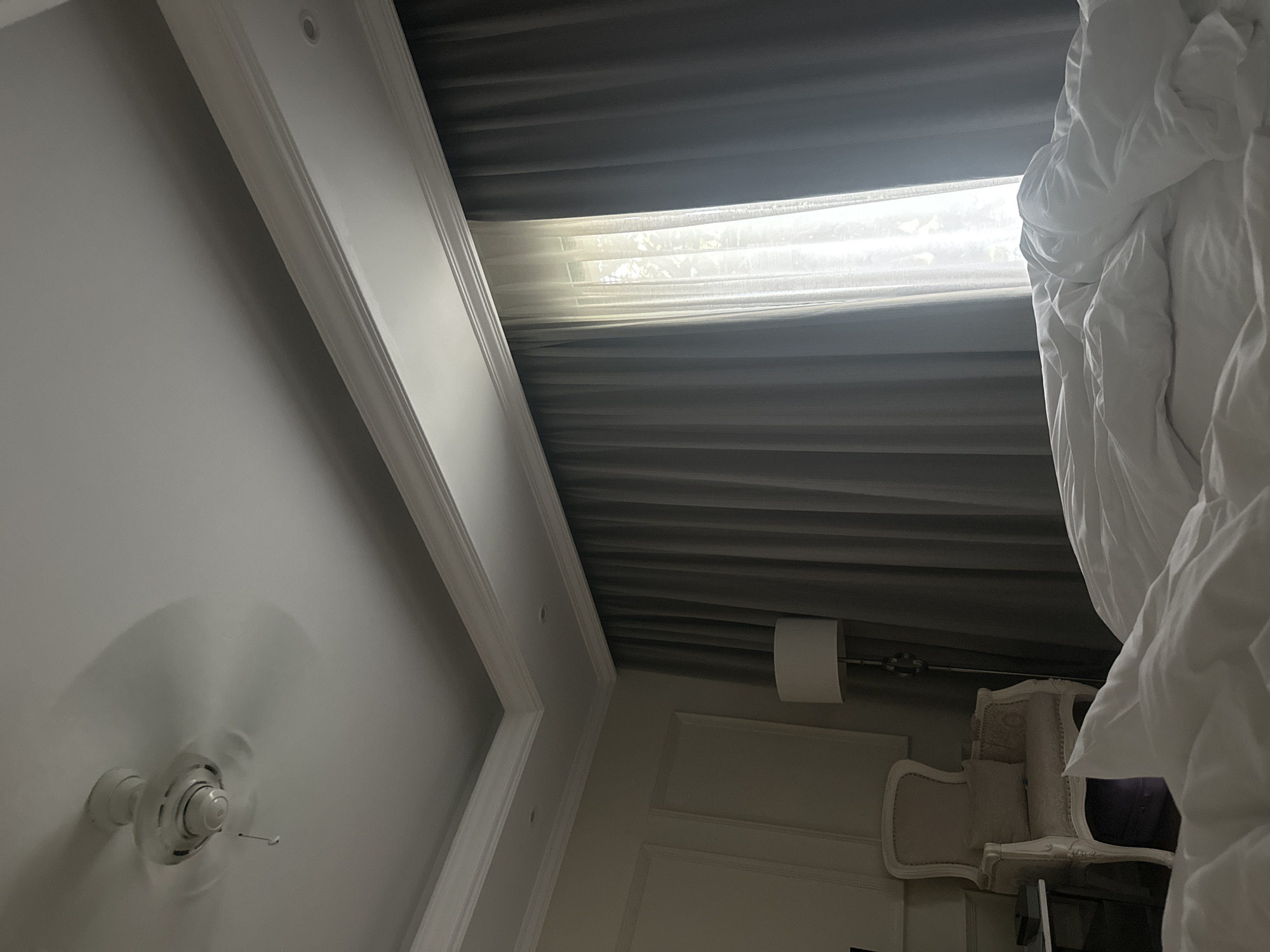 This rotated indoor photograph captures a bright, comfortable room, likely a bedroom, viewed from a low angle. In the lower left, a white ceiling fan with blurred blades is in motion against a light-colored ceiling. White crown molding runs along the ceiling and walls, featuring recessed lighting.
A large window dominates the upper center, partially covered by drawn dark gray drapes. Bright natural light streams through a sheer white curtain visible between the drapes, hinting at outdoor foliage beyond. In the foreground on the right, a crumpled white duvet or bedspread is visible, suggesting a bed is nearby.
Towards the mid-right, partially obscured by the duvet, stands a classic-style armchair with light-colored upholstery and intricate carvings. A white drum-shaped lampshade is mounted on a fixture near the window, and the wall behind the chair has decorative white paneling. The bright light from the window clearly indicates it is daytime. No specific activity is depicted, and no text is visible.