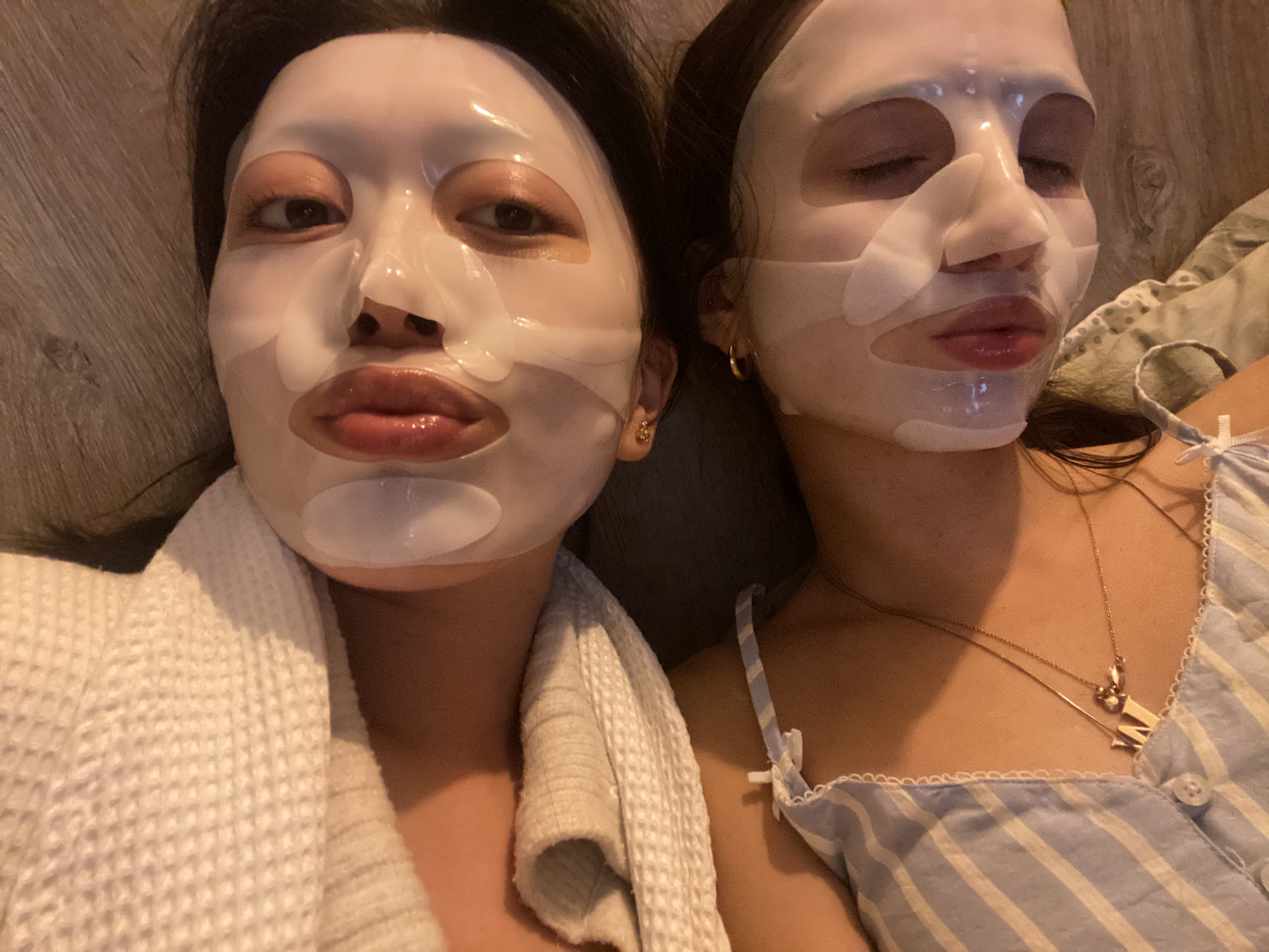 Two young individuals, likely women, are captured in a close-up, high-angle shot, seemingly lying down next to each other in what appears to be a cozy indoor setting. Both are engaged in a self-care routine, wearing white sheet face masks that cover most of their faces, leaving cutouts for their eyes, nostrils, and mouths.
The person on the left has dark hair and one eye open, looking directly at the camera, while their lips, visible through the mask's opening, are glossy. They are wearing a white, textured robe or towel, partially visible around their neck and shoulder. A small gold earring is visible in their left ear.
The individual on the right has dark hair and appears more relaxed, with their eyes closed or mostly closed. Their lips are also visible and slightly parted. They are wearing a light blue and white striped top with thin straps and a lacy trim. A delicate gold necklace with two charms, one resembling the letter 'M', rests on their chest. A gold hoop earring is visible in their right ear.
The background features a warm-toned wooden surface, possibly flooring or a wall, adding to the home-like ambiance. The lighting is soft and dim, suggesting the activity is taking place in the evening or at night. The overall impression is one of shared relaxation and comfort. No visible text is present in the image.