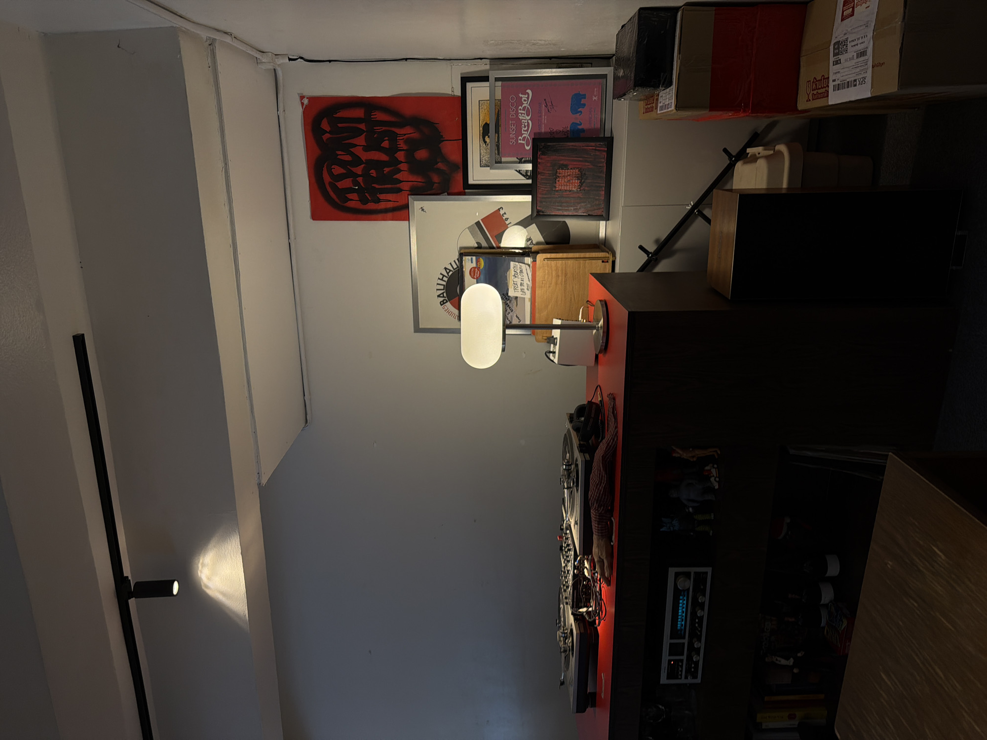This image depicts a creatively arranged collection of items in what appears to be a domestic or studio setting. The scene is dominated by various framed art pieces and records displayed on a wall, suggesting a passion for music and art. Among the artworks, a red banner with black graffiti-style lettering is prominent. Another framed piece features the text "SUNSET DISCO Break Bot" and an illustration with elephants. Below these, records with album art, including one with "CHES BAUHAU" and "1923" visible, are stacked and leaning against the wall.
A modern desk lamp with a white, elongated shade is positioned in front of the artwork, casting a soft light. The overall impression is one of a personalized space, possibly a living room or a creative workspace, filled with items that hold personal significance. The presence of turntables and audio equipment further reinforces the theme of music. Boxes are stacked on shelves in the background, and some are taped shut, possibly indicating storage or recent acquisitions. The lighting in the room is a mix of ambient light and focused light from the lamp, creating some shadows and highlighting specific areas. The orientation of the image is such that the scene appears vertical. There are no people visible in the frame. The location context of Chiang Mai City Municipality, Thailand, does not provide specific visual cues within the image itself.