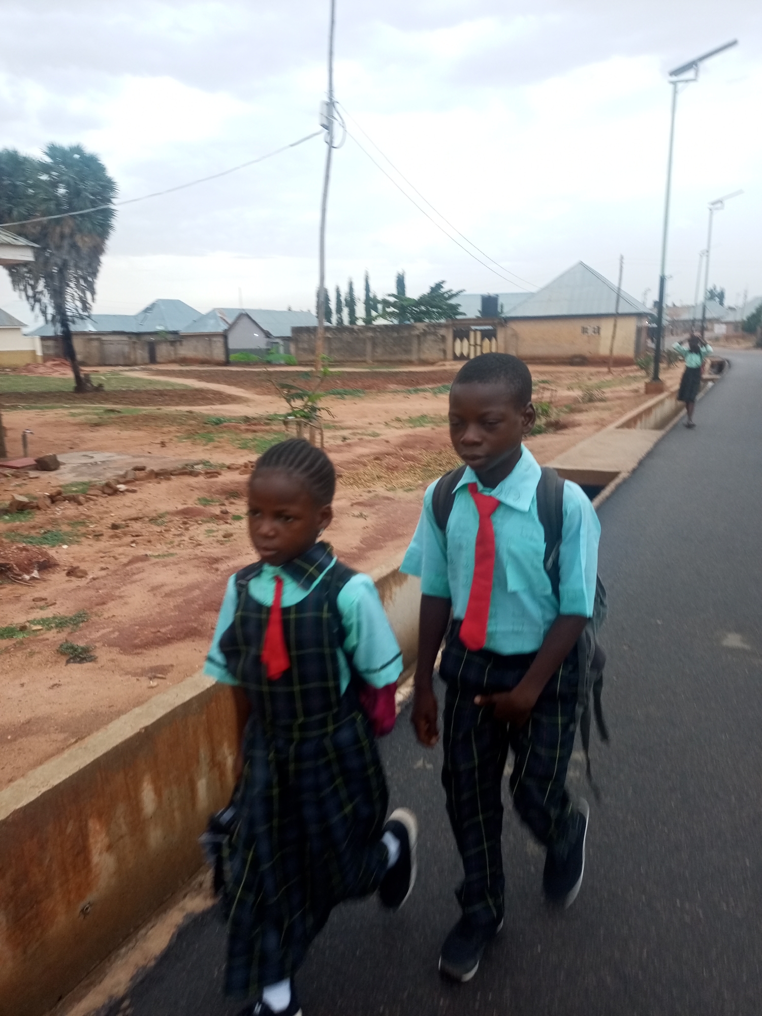 Two children in school uniforms walk along a paved road. The girl on the left wears a plaid dress over a light blue shirt with a red tie and black shoes. The boy on the right wears a light blue short-sleeved shirt, plaid pants, black shoes, a red tie, and a backpack. The location is Gombe, Nigeria. The background shows houses, a dirt area, and utility poles. Another person walks further down the road. The sky is overcast. A concrete curb separates the road from the unpaved area.