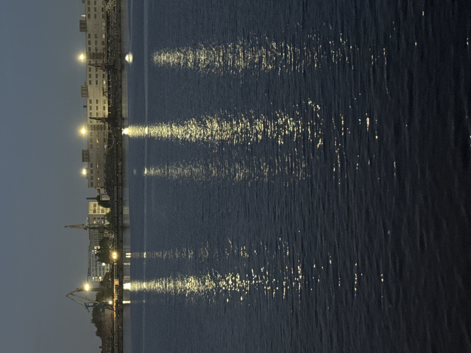 The image captures a nocturnal view of a waterfront industrial or urban area. On the left, a shoreline is lined with several multi-story buildings, many displaying illuminated windows. Interspersed along the bank are numerous large cranes, some with distinct yellow and red components, suggesting the presence of a port or shipyard. Artificial lights from these structures and possibly streetlights cast their glow across the scene.
To the right, a dark body of water, likely a river or bay, dominates the foreground and midground. The water's surface is relatively calm, allowing for prominent reflections of the onshore lights. These reflections appear as shimmering, elongated streaks of bright yellow and white light, extending horizontally across the dark water and creating a striking luminous pattern. The sky above is dark, confirming the time of day as night or late dusk. There are no visible people or discernible text in the photograph. The overall atmosphere is one of quiet, nocturnal activity or calm.