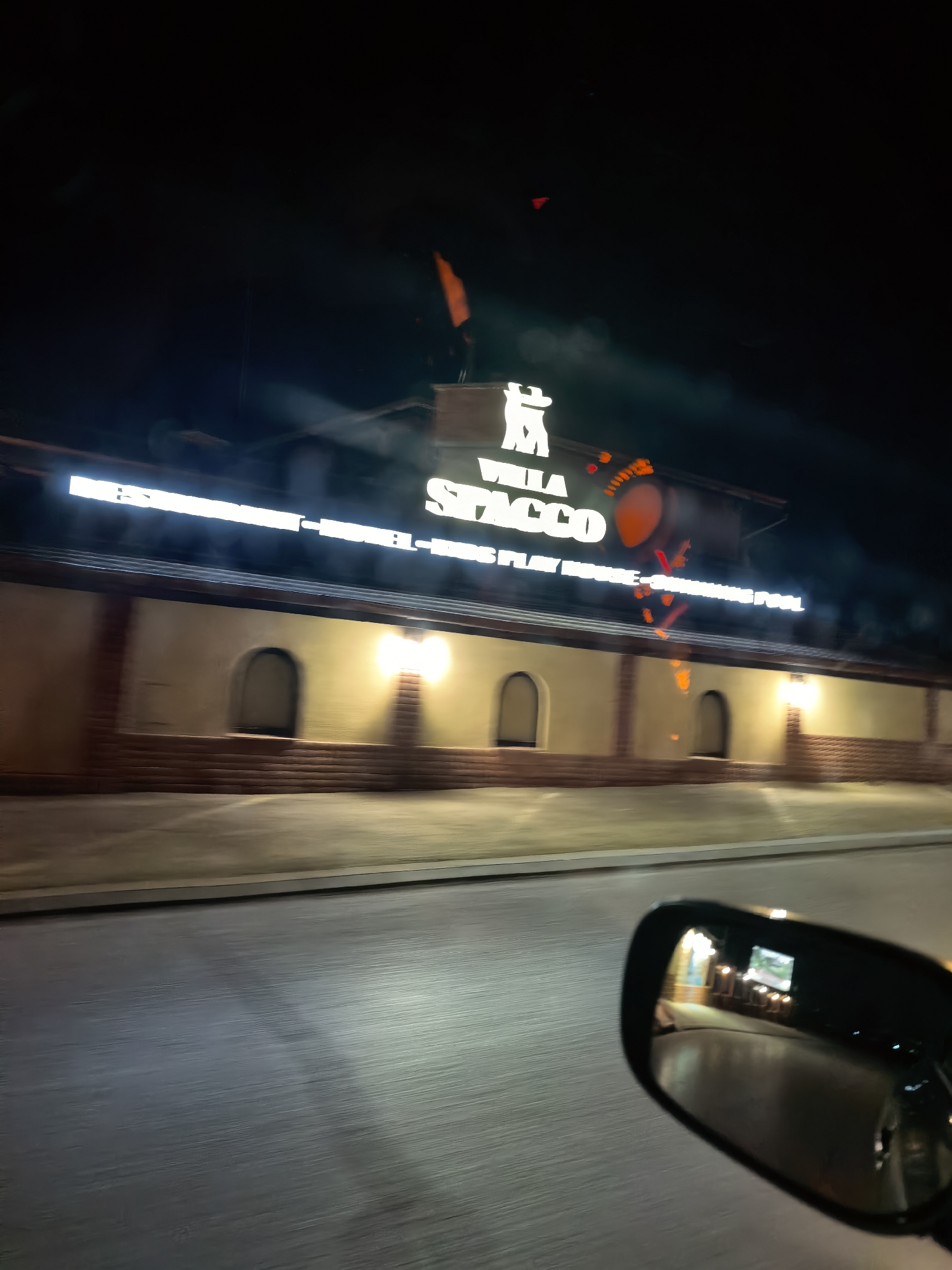 At night in an urban environment, a building with illuminated signage reading "Villa Stacco" and "Play House - Swimming Pool" is visible. The building has arched windows or doorways and is illuminated by exterior lights. The scene is captured from a moving vehicle, as evidenced by the side mirror reflecting other lights. The street appears wet, reflecting light.