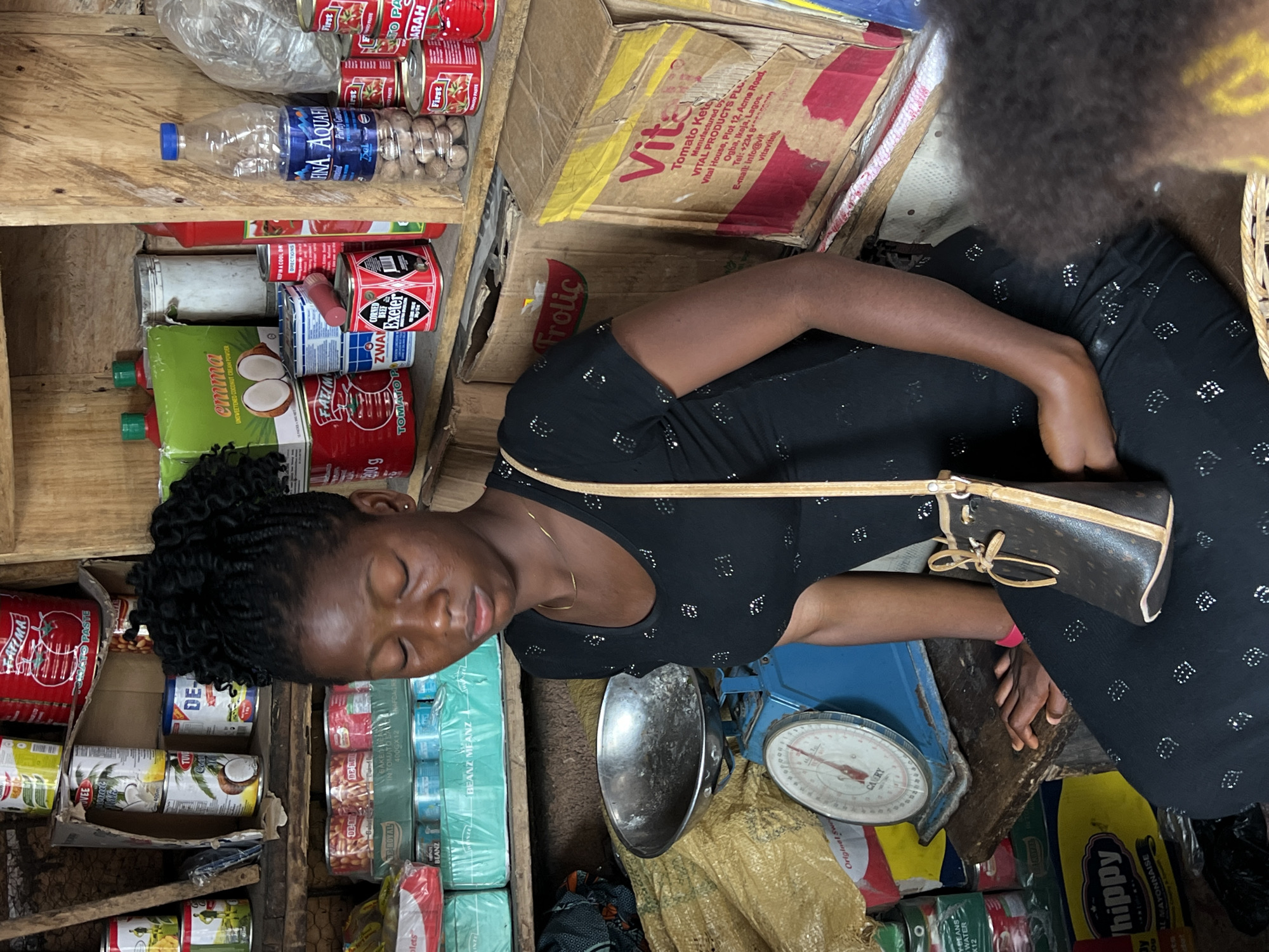 In Nkpor, Nigeria, a young Black woman is captured in a market stall, her eyes closed in what appears to be a moment of rest or quiet contemplation. She wears a black dress adorned with small, reflective diamond-shaped patterns. A simple gold chain necklace rests against her collarbone, and a small, dark brown drawstring bag with light brown trim is slung over her shoulder.
The background is a dense arrangement of shelves overflowing with various consumer goods. Cans of tomato paste, coconut milk, and what appear to be beans are stacked prominently. Brands such as "Fatima," "Tuvée," "Emma," and "First" are visible on the packaging. A large blue mechanical scale sits to the left of the woman, topped with a metal bowl. Behind her, more shelves are laden with bottles of water, nuts, and other packaged foods. The overall environment suggests a bustling marketplace, with the woman positioned in a central, yet somewhat secluded, spot amidst the commercial offerings.
